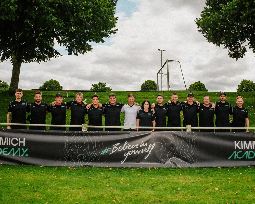 Group of athletic people in black sports shirts standing outdoors behind a black banner with white text on a grassy field with trees and a partly cloudy sky in the background.