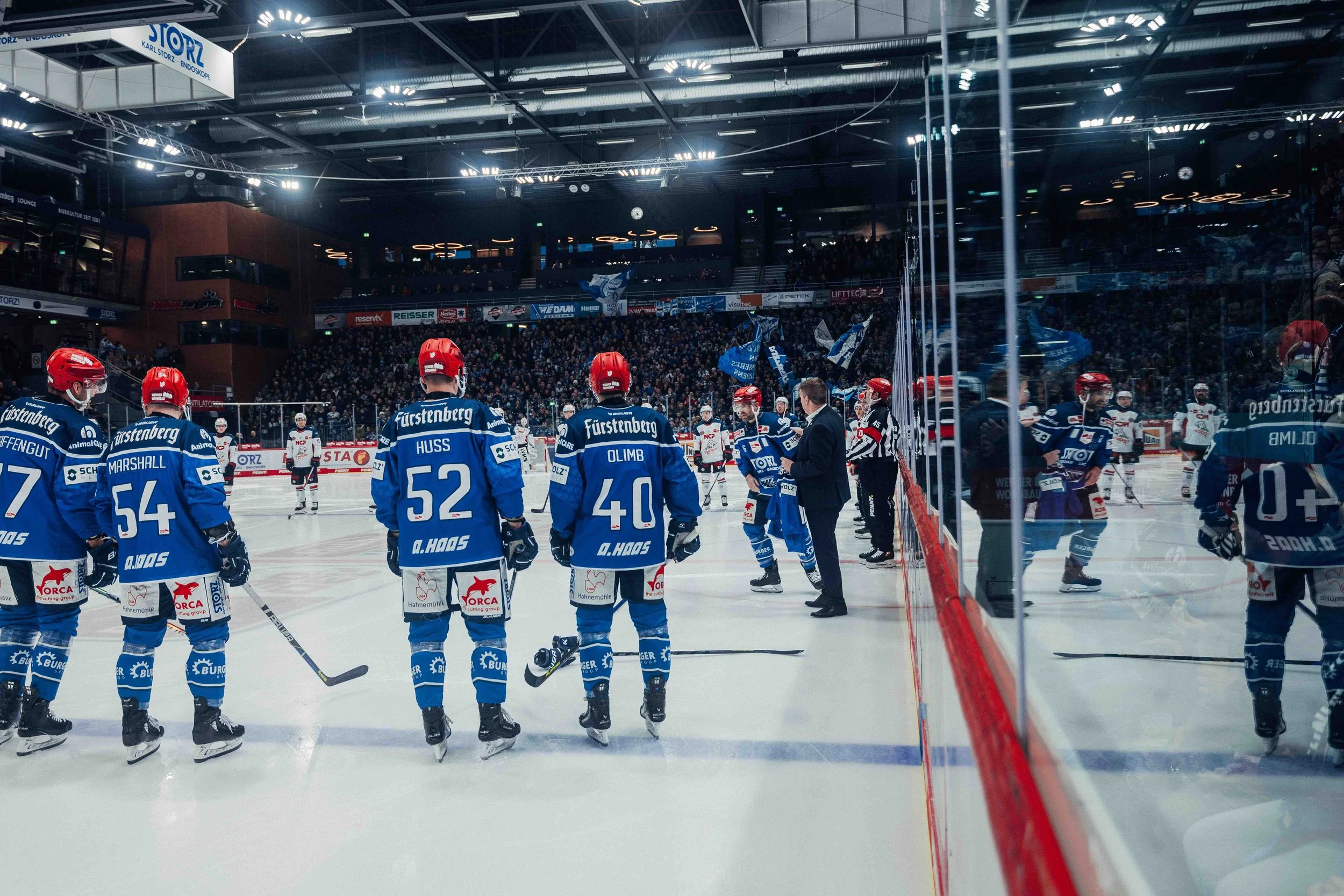 Ice hockey players in blue uniforms lining up on the ice rink during a game, with spectators watching from the stands in the background.