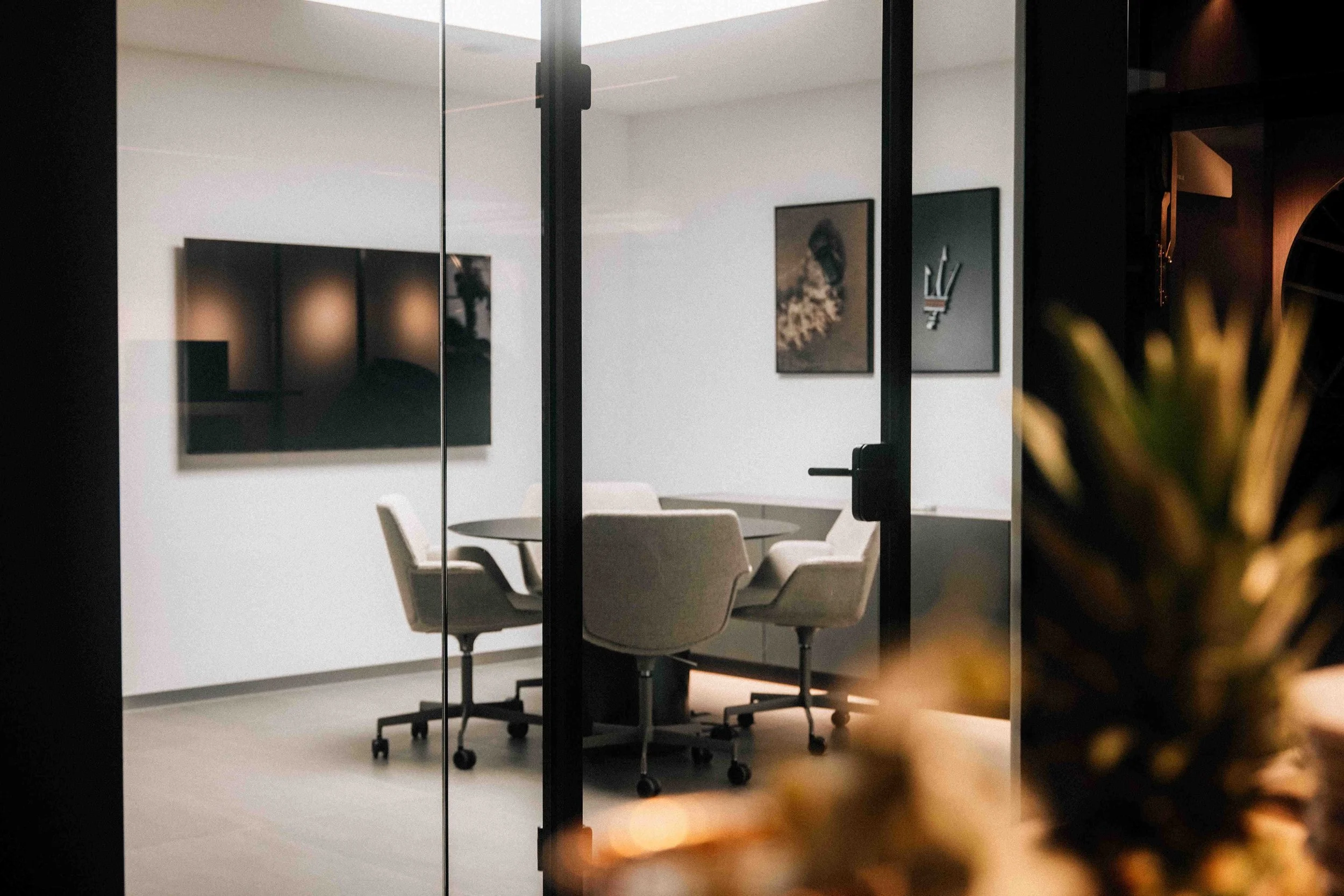 A modern conference room viewed through glass walls, with a round table and four beige chairs on casters, and framed artwork on white walls.