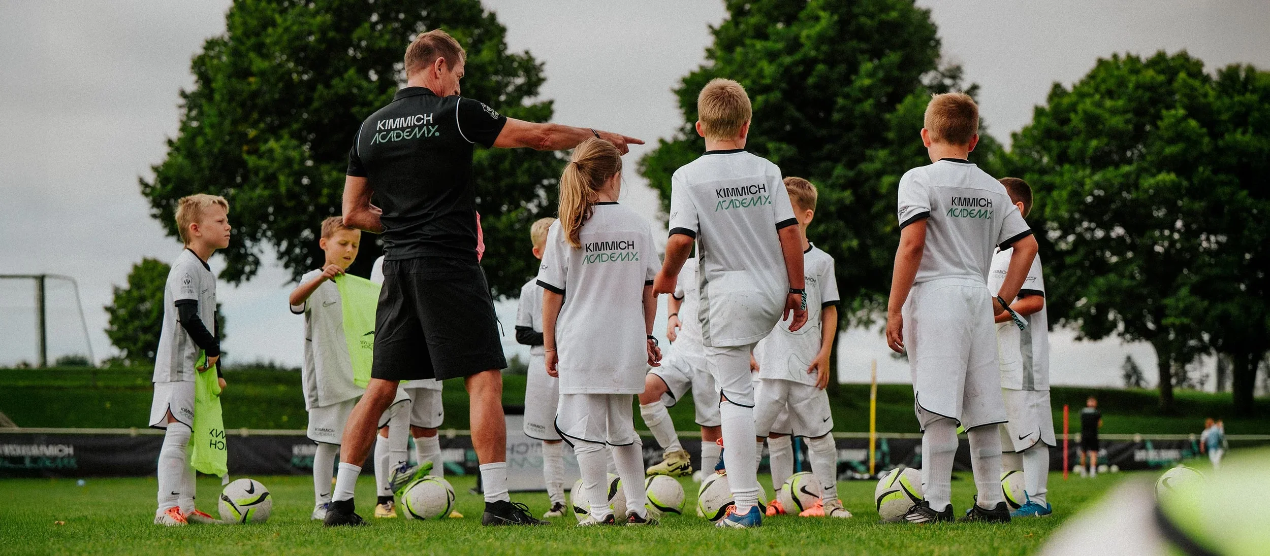 Children in white soccer uniforms listening to coach during practice on a green field with trees in the background.