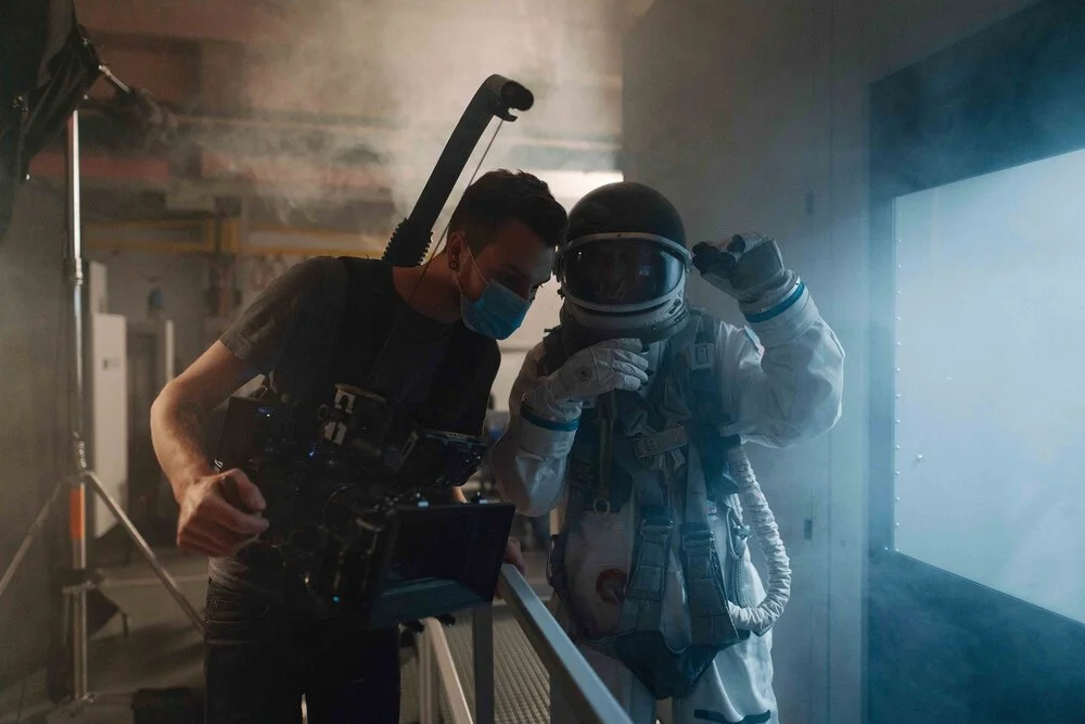 A film director and an astronaut are on a film set, with the director wearing a face mask and operating a camera while the astronaut is dressed in a spacesuit, adjusting their helmet in a smoky room with a large screen.