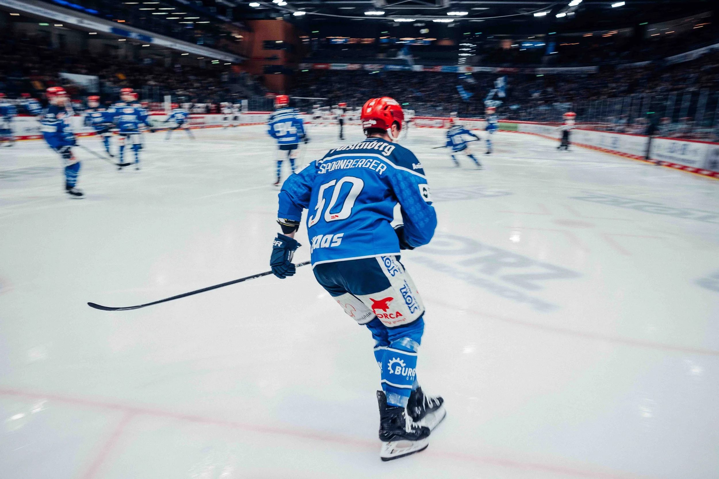 A hockey game in progress with players on the ice, focused on a player wearing a blue jersey , a red helmet, and holding a hockey stick, skating across the rink.