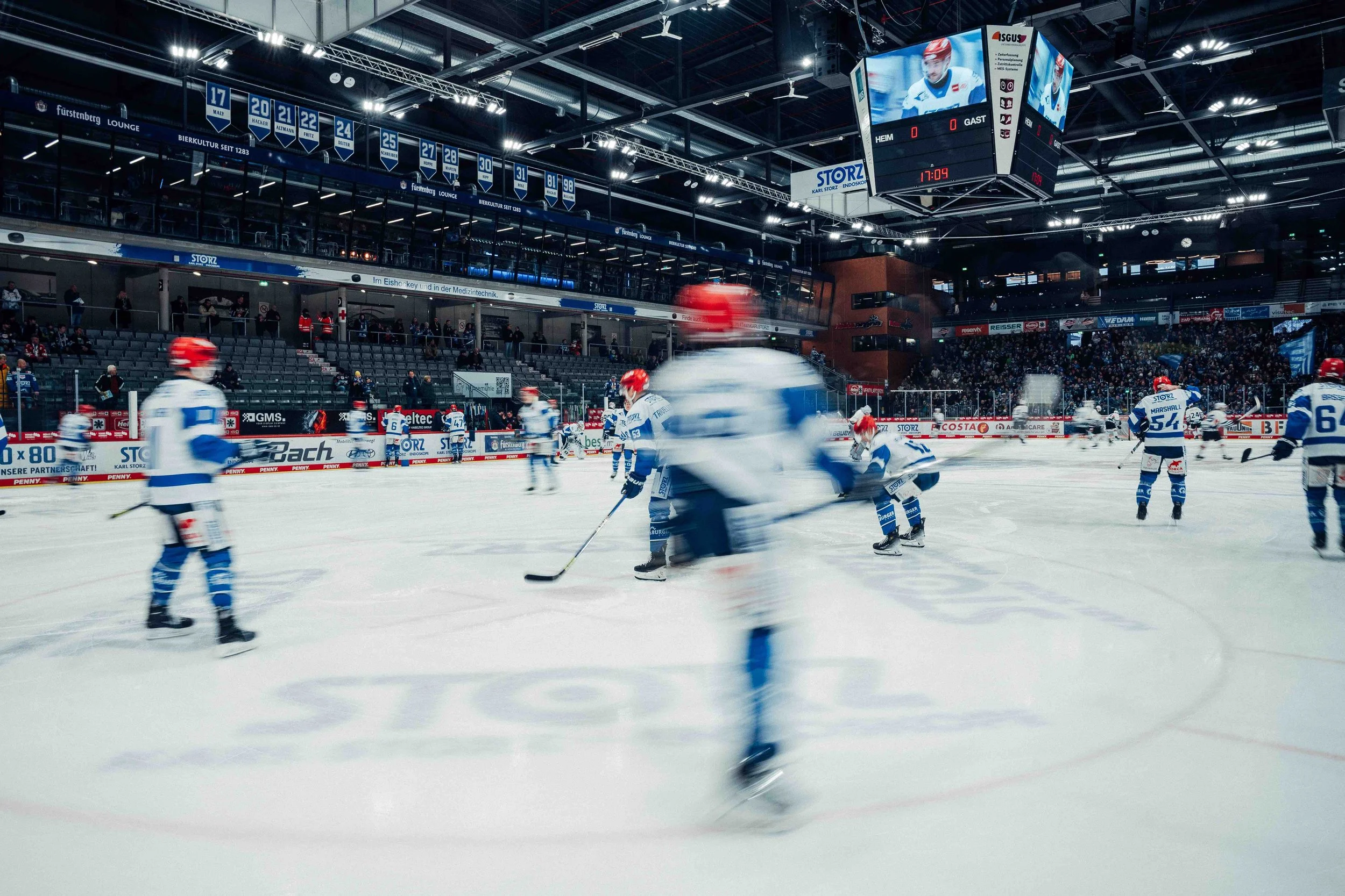 Ice hockey game in progress at an indoor arena, players in white and blue uniforms, spectators in the stands, large scoreboard hanging from ceiling showing the game time and player stats.