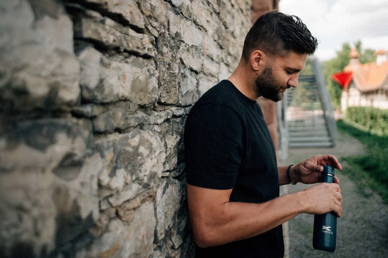 A man in a black shirt stands next to a stone wall, holding a dark water bottle and looking into it.