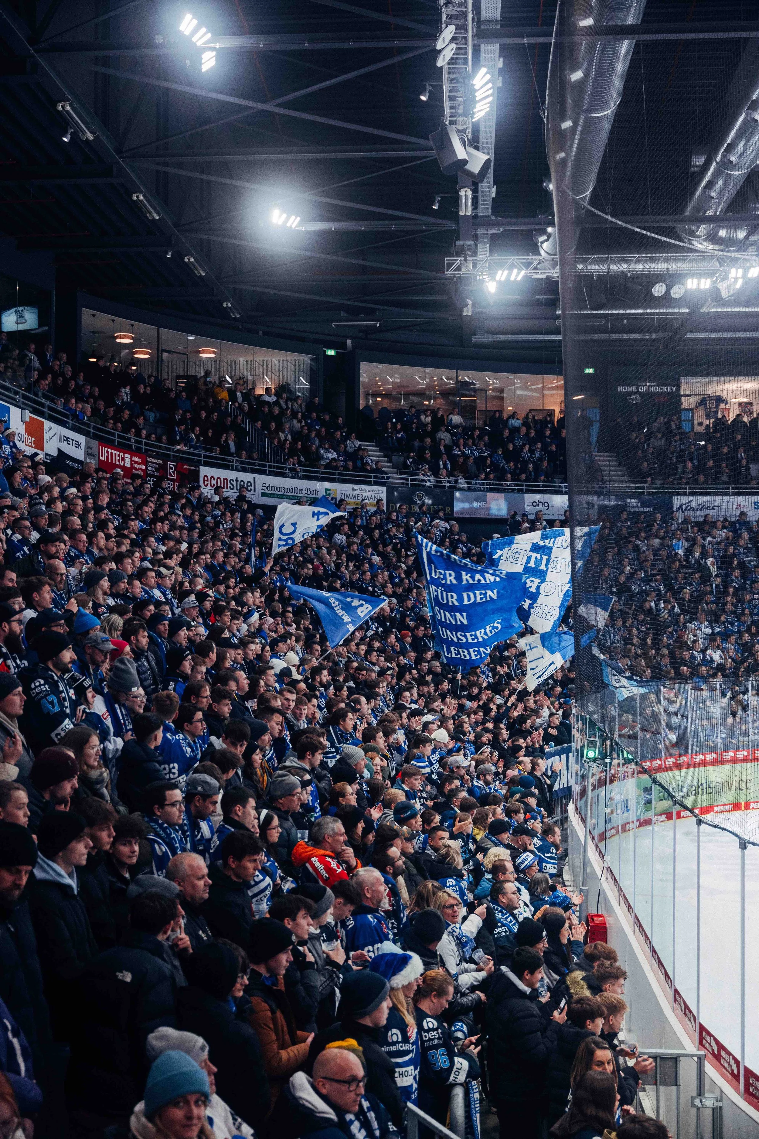 Crowd of hockey fans in an ice rink, many wearing blue and white jerseys, waving flags, and supporting their team.