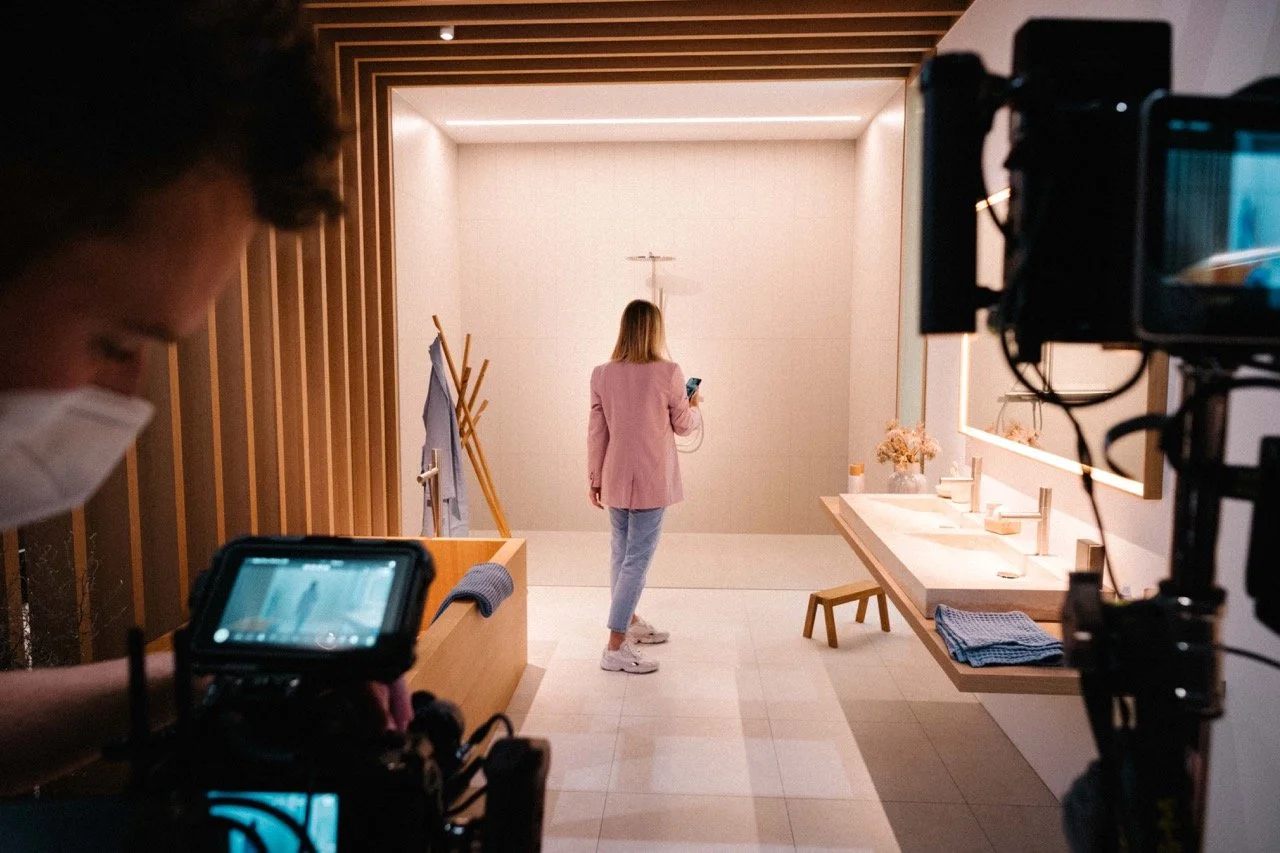 A woman standing in a modern, well-lit bathroom, looking at her phone. The bathroom has a long mirror, a small stool, a towel on a rack, and minimalist decor with flowers on the counter. A person with a mask and camera is filming or photographing the scene.