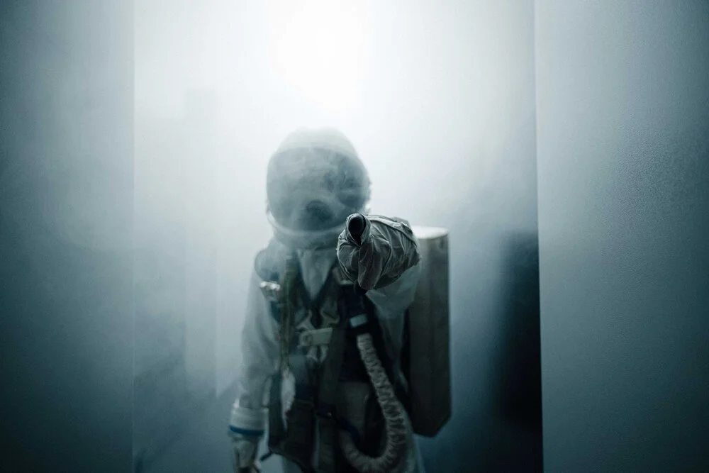 A person in astronaut gear, including a helmet and mask, pointing a gun or spray nozzle in a narrow, foggy corridor, with a backpack or tank on their back.
