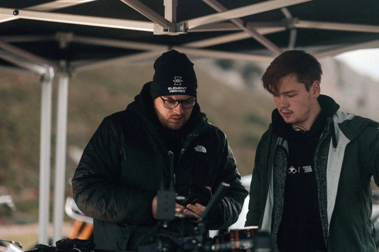 Two men standing under a canopy, looking at a device, outside during daytime