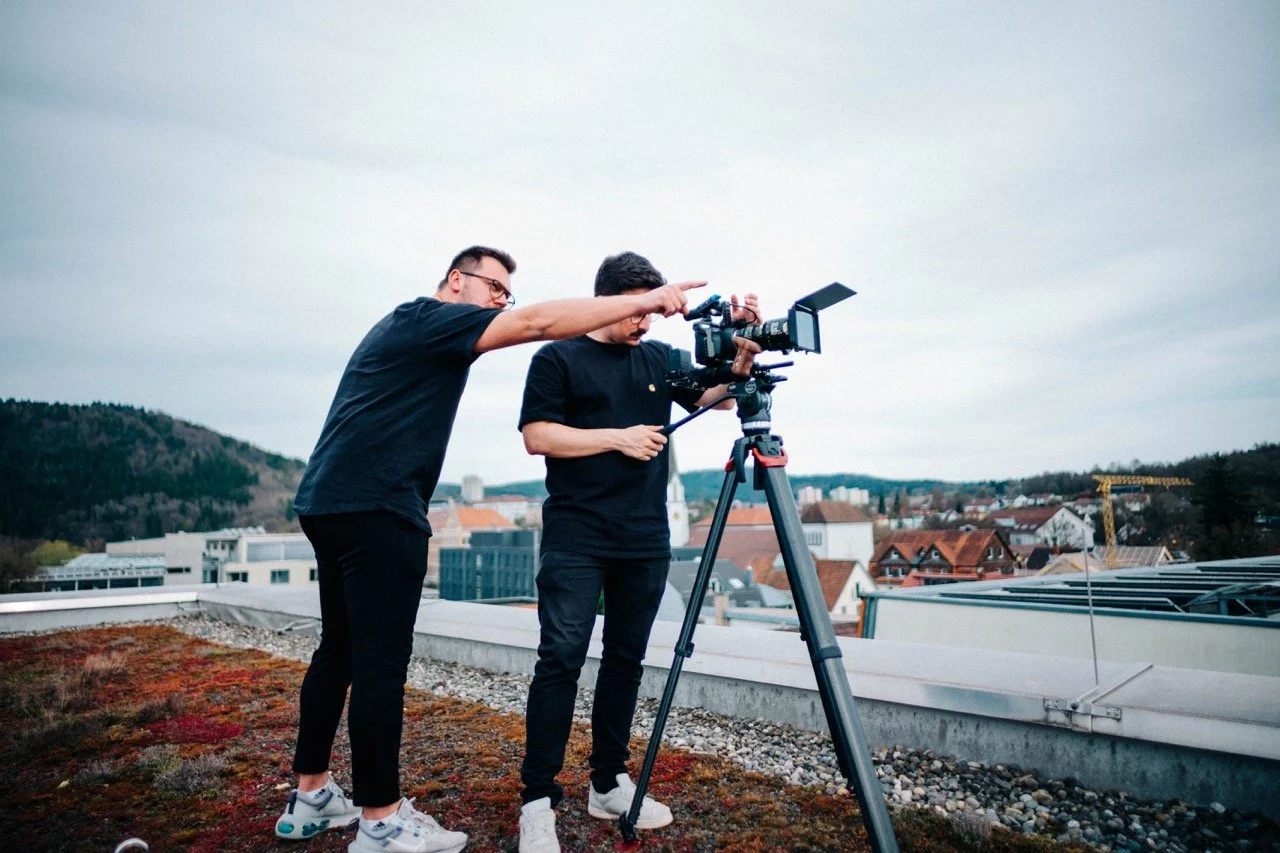 Two men setting up a video camera on a rooftop overlooking a cityscape with a crane in the background.