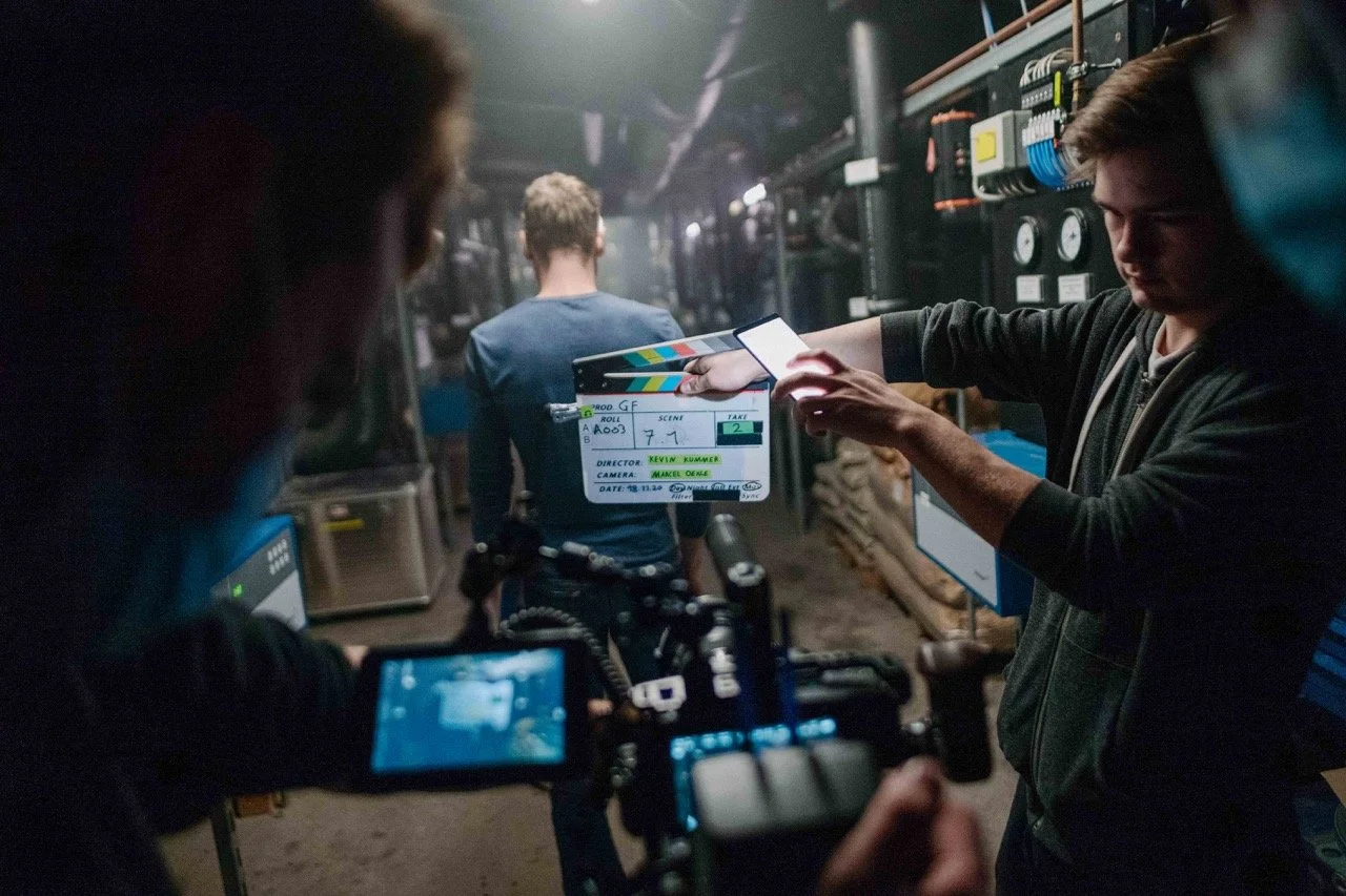 Film crew on set with a clapperboard, camera operator, and a man in the background in a dimly lit industrial or mechanical setting.