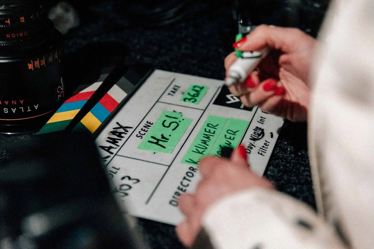 A person with red nail polish writing on a film clapperboard for a production titled 'Mr. S! S.' with scene number '354A' and additional notes, surrounded by camera lenses.