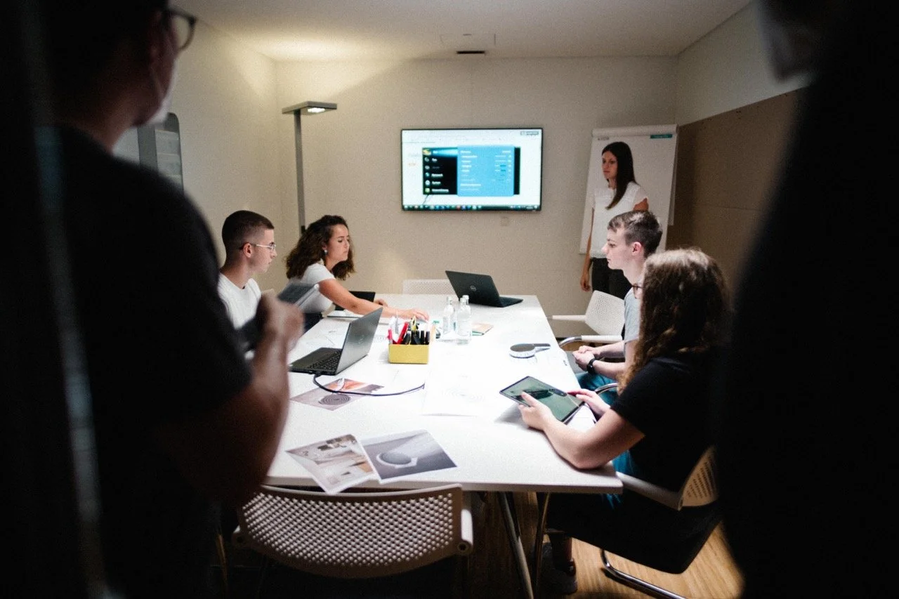 A group of six young adults are gathered in a modern conference room, engaged in a meeting with laptops, tablets, and documents. One woman is standing in front of a flip chart, presenting to the group, while a large screen on the wall displays a computer interface. The room has minimal decor and is well-lit.