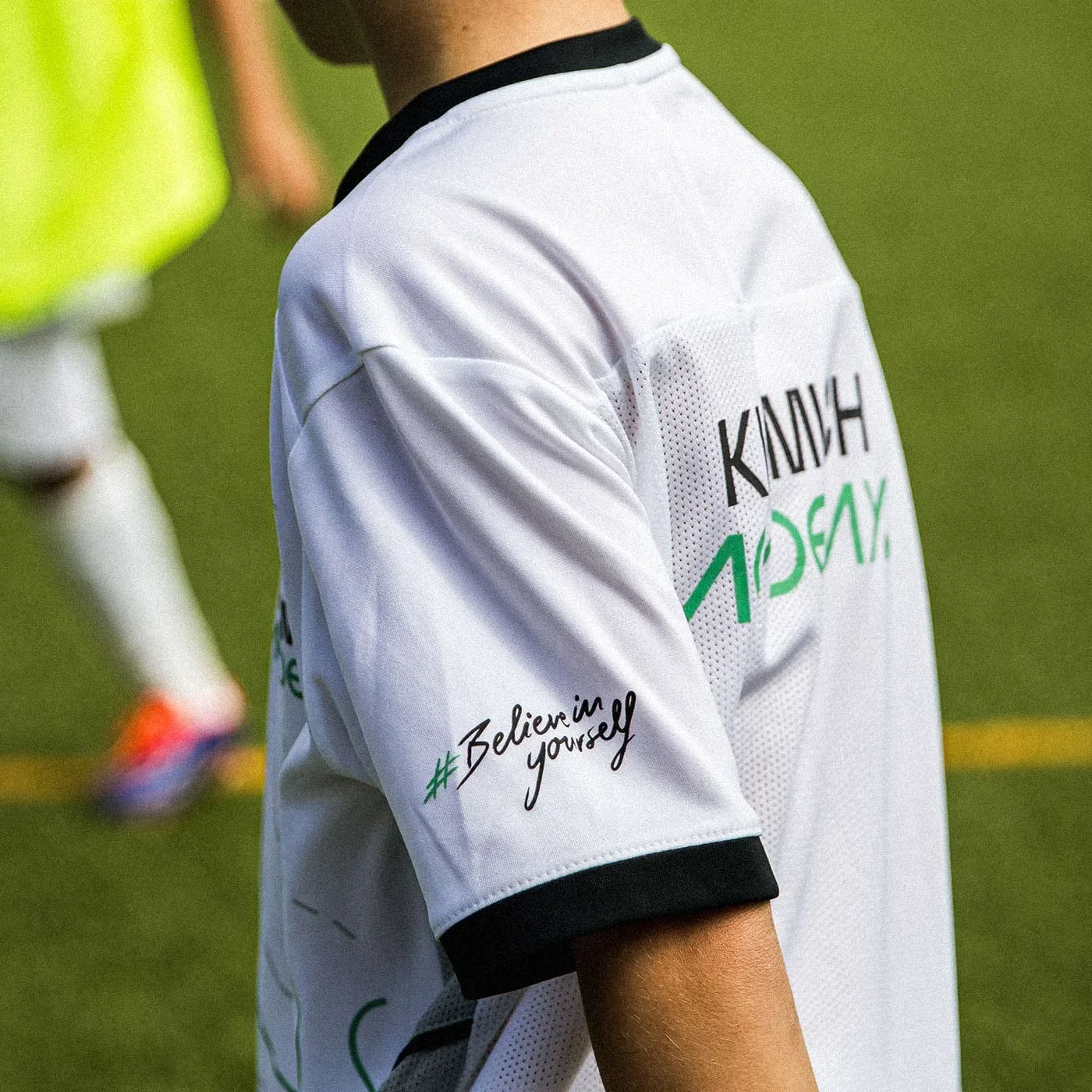 Close-up of a person wearing a white sports jersey with black trim, featuring the hashtags #BelieveYourself and a large graphic text on the back. The background shows a blurred green field and part of another person.