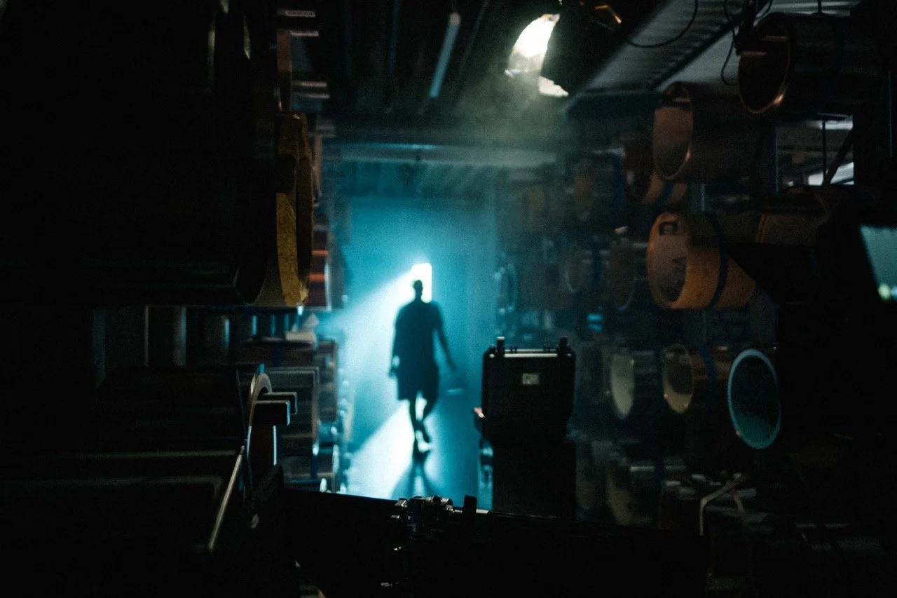 Silhouette of a person walking down a dark aisle illuminated by a bright light from behind, with shelves filled with equipment on either side.