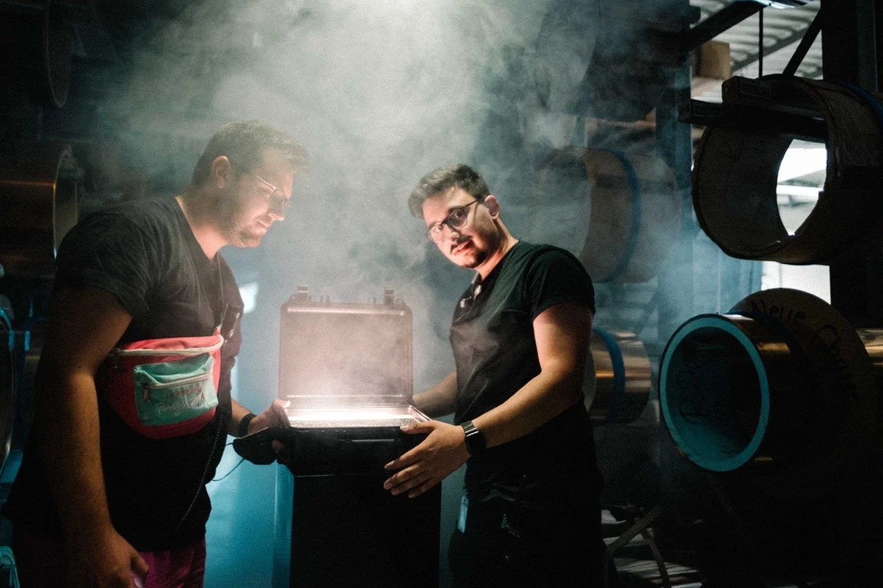 Two men in sunglasses and black shirts looking at a glowing case in a dark, smoky room filled with equipment and equipment racks.