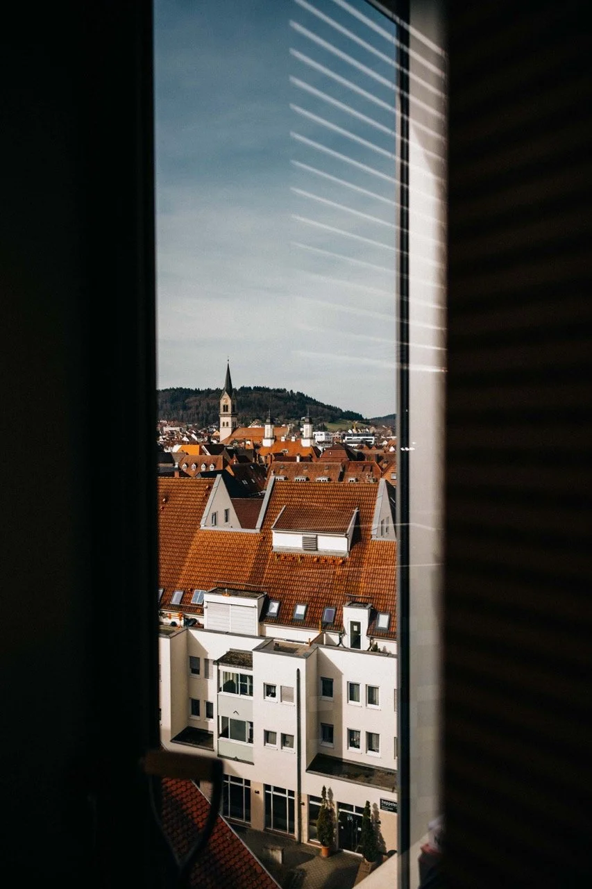 View through a window with blinds showing a cityscape with orange-roofed buildings, a church steeple, and a hilly landscape in the background.