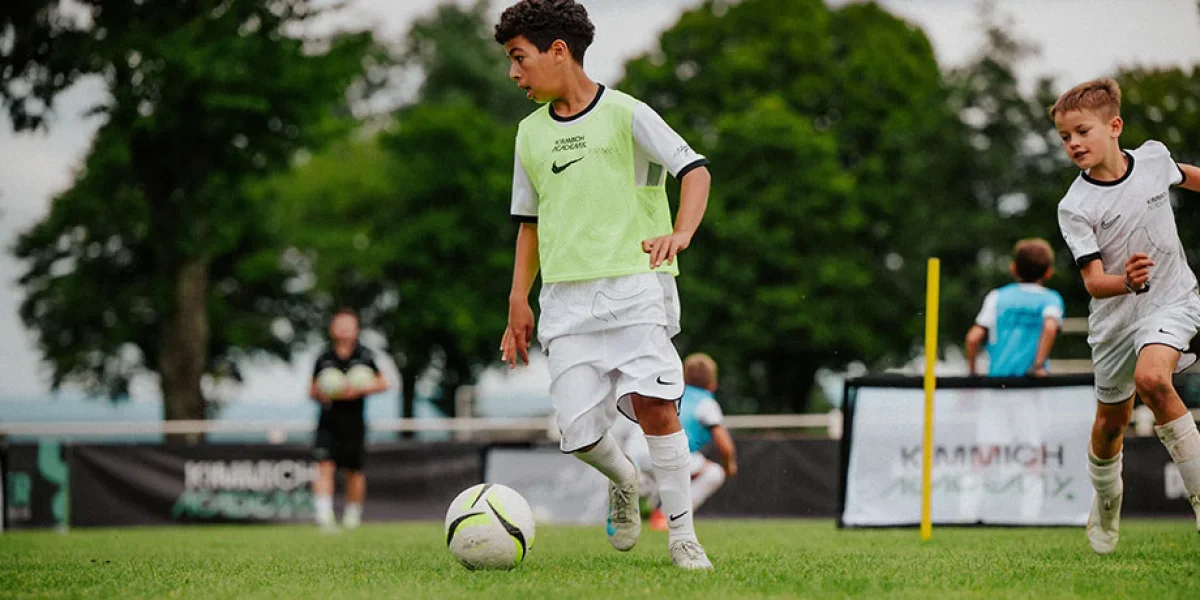 Young boys playing soccer on a field during daytime, with trees and soccer goal in background.