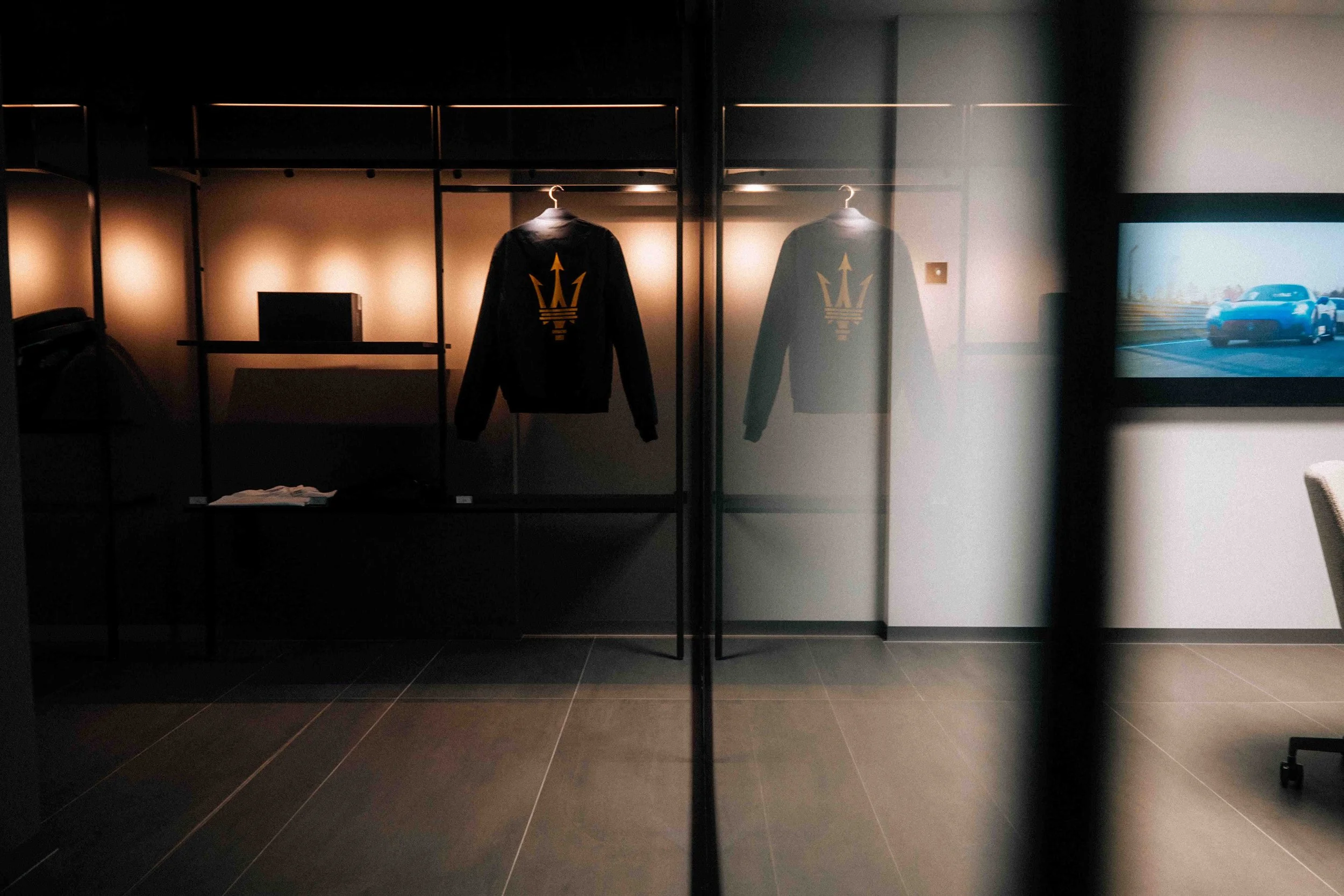 A display case with two black jackets featuring a gold trident logo, viewed through a glass wall in a modern room with a television showing a blue car on a racetrack.