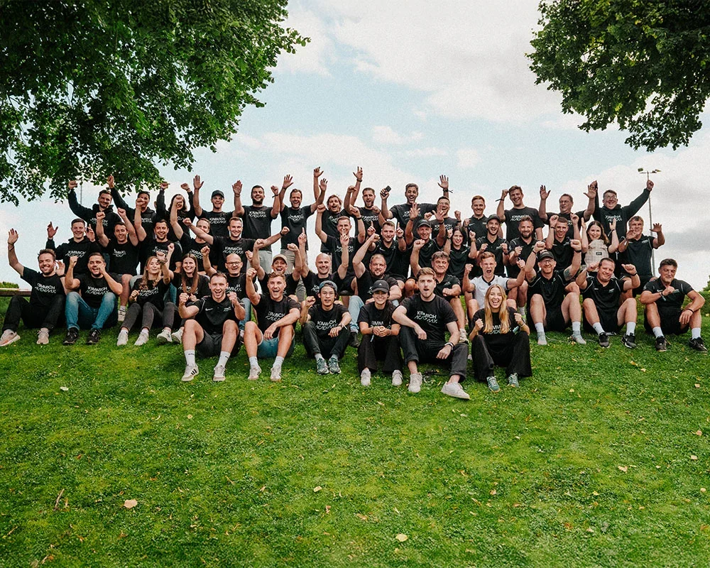 Group of people outdoors on a grassy hill, smiling and celebrating with raised hands, wearing matching black shirts.
