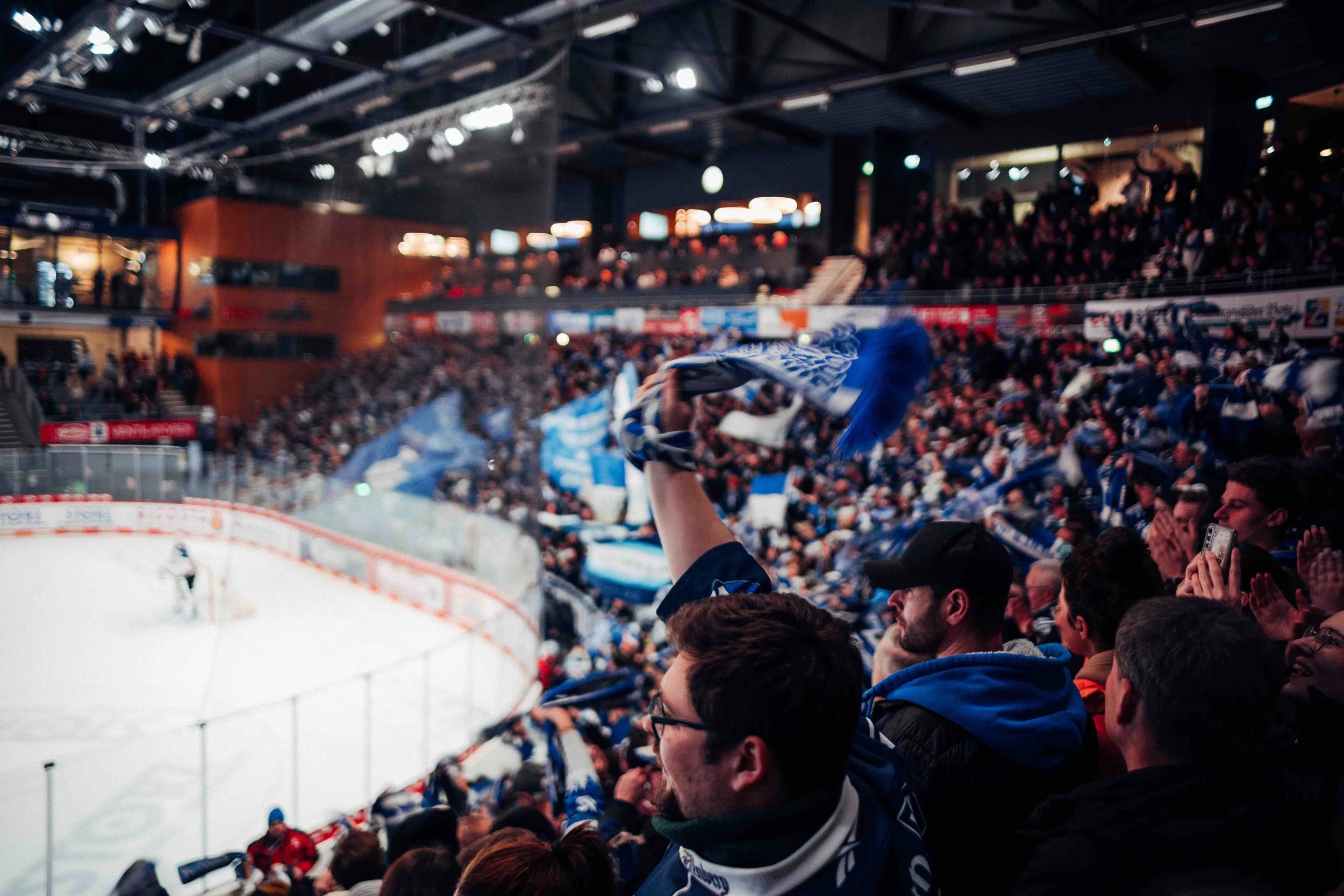 A crowd of hockey fans in an indoor arena, cheering and waving scarves as a game is played on the ice rink.