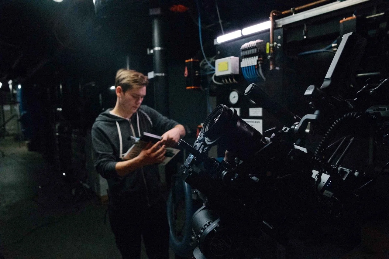 A young man in a black hoodie examining a book or manual near a large professional camera or telescope in a dark room with control equipment on the wall.