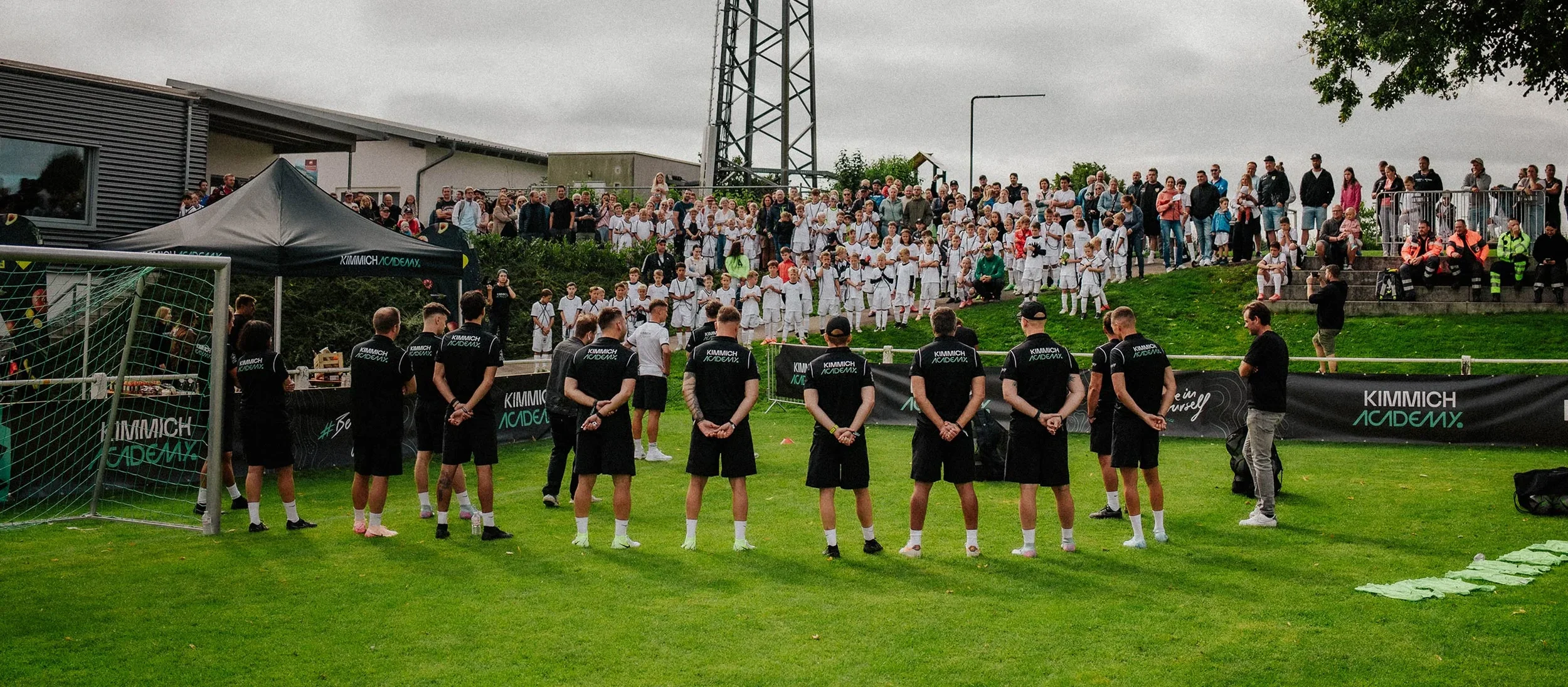 Youth soccer team standing on the field during an outdoor event, with a crowd of spectators and participants in karate uniforms in the background on a cloudy day.