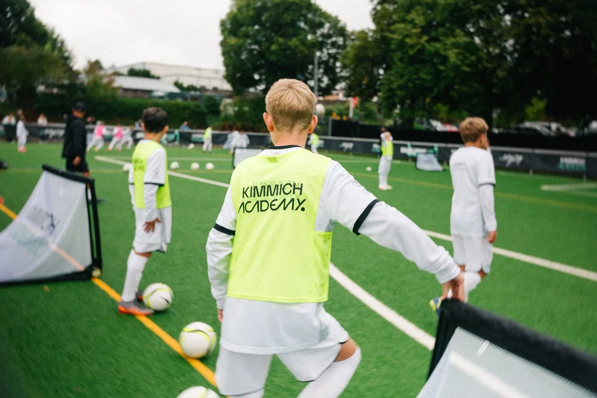 Children in soccer uniforms and training bibs on a field during practice, with soccer balls and cones visible.