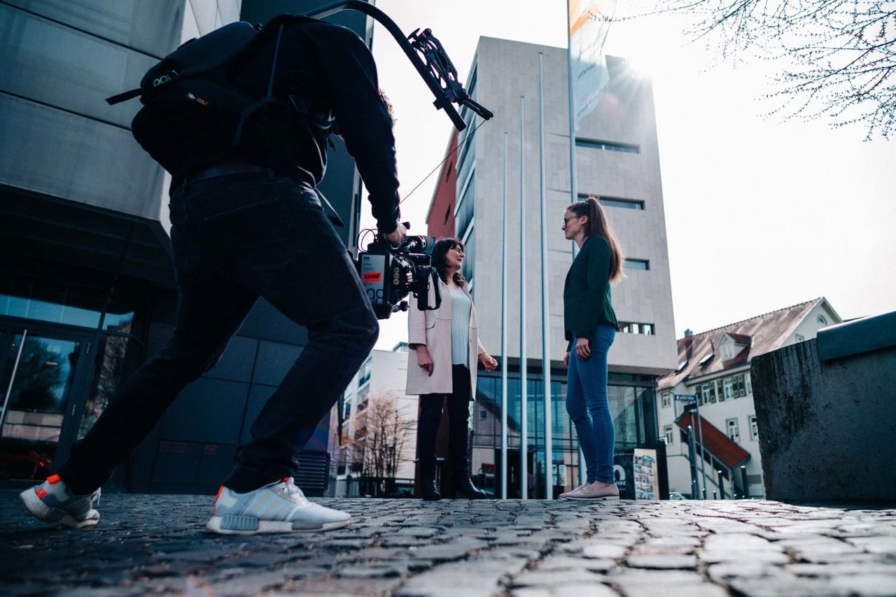 A person filming two women having a conversation outdoors in front of a modern building.