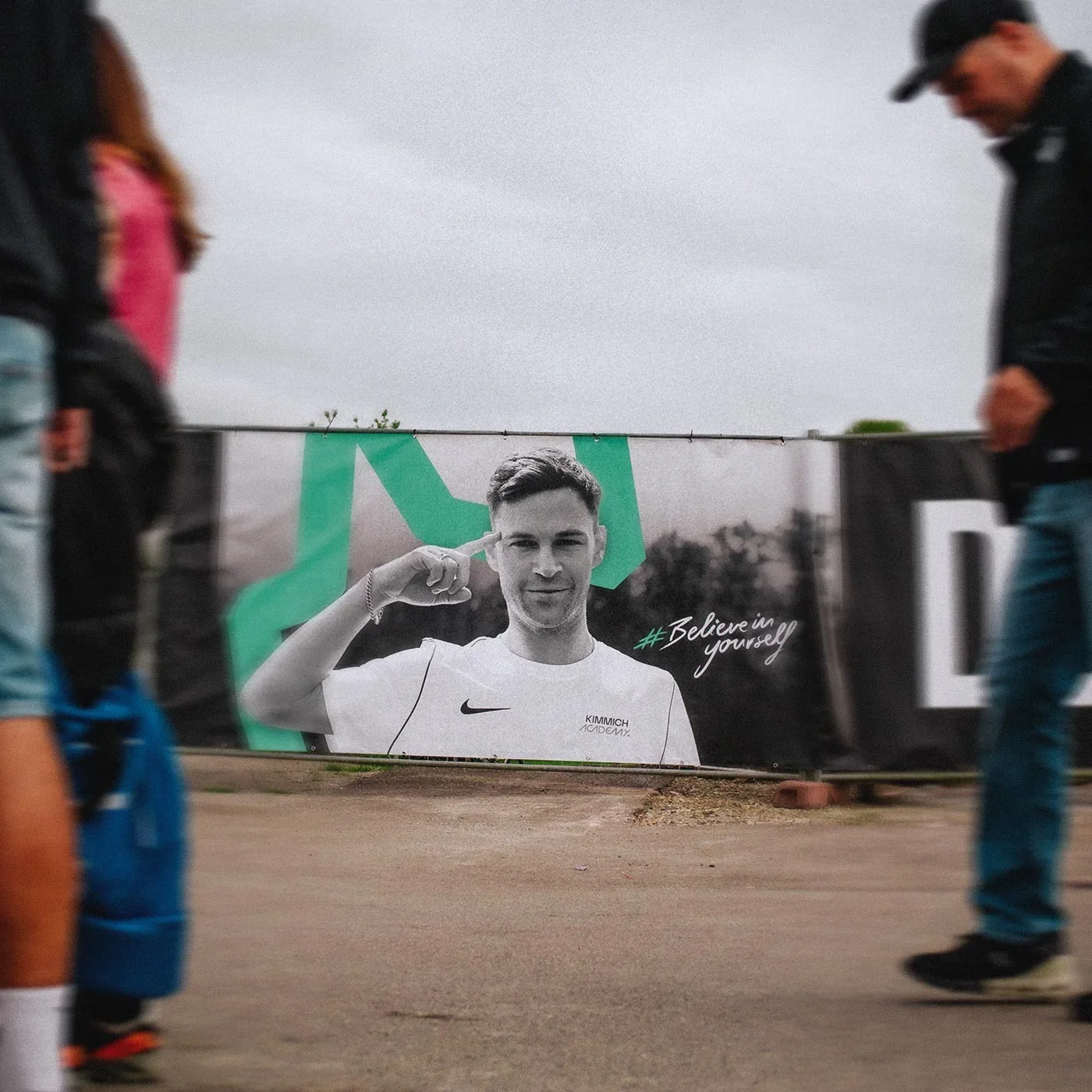 A large advertisement banner featuring a young man in a white athletic shirt with a Nike logo, giving a thumbs-up and smiling. The banner has the hashtag #Believeinyourself and is black, white, and green. Several people are walking past the banner ou
