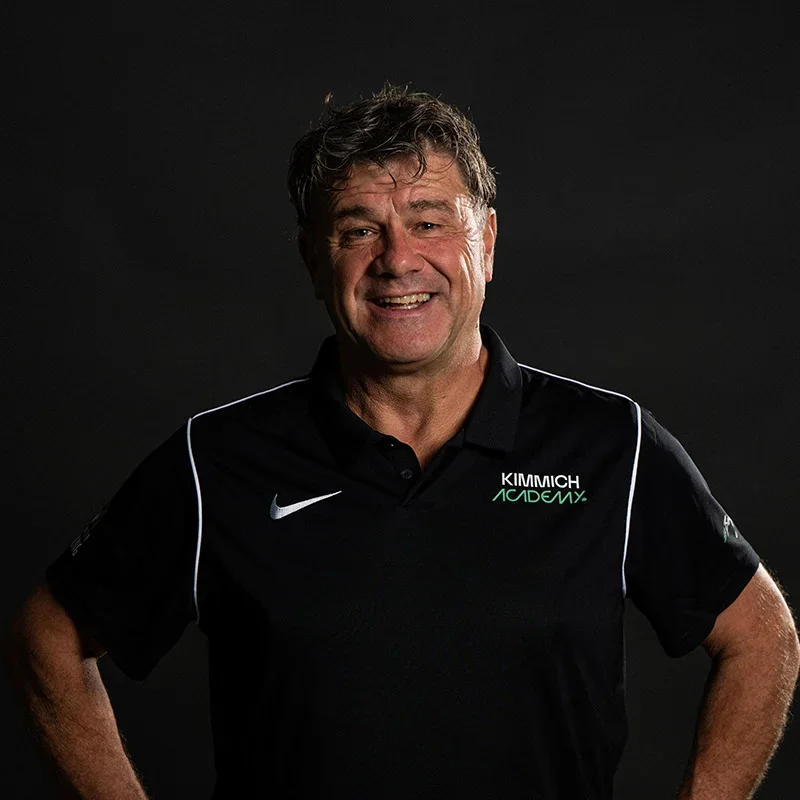 A smiling man wearing a black Kimmich Academy polo shirt with white accents and a Nike logo, standing against a dark background.