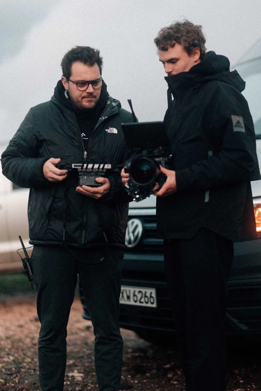Two men filming with professional camera equipment outdoors, with a Volkswagen van in the background.