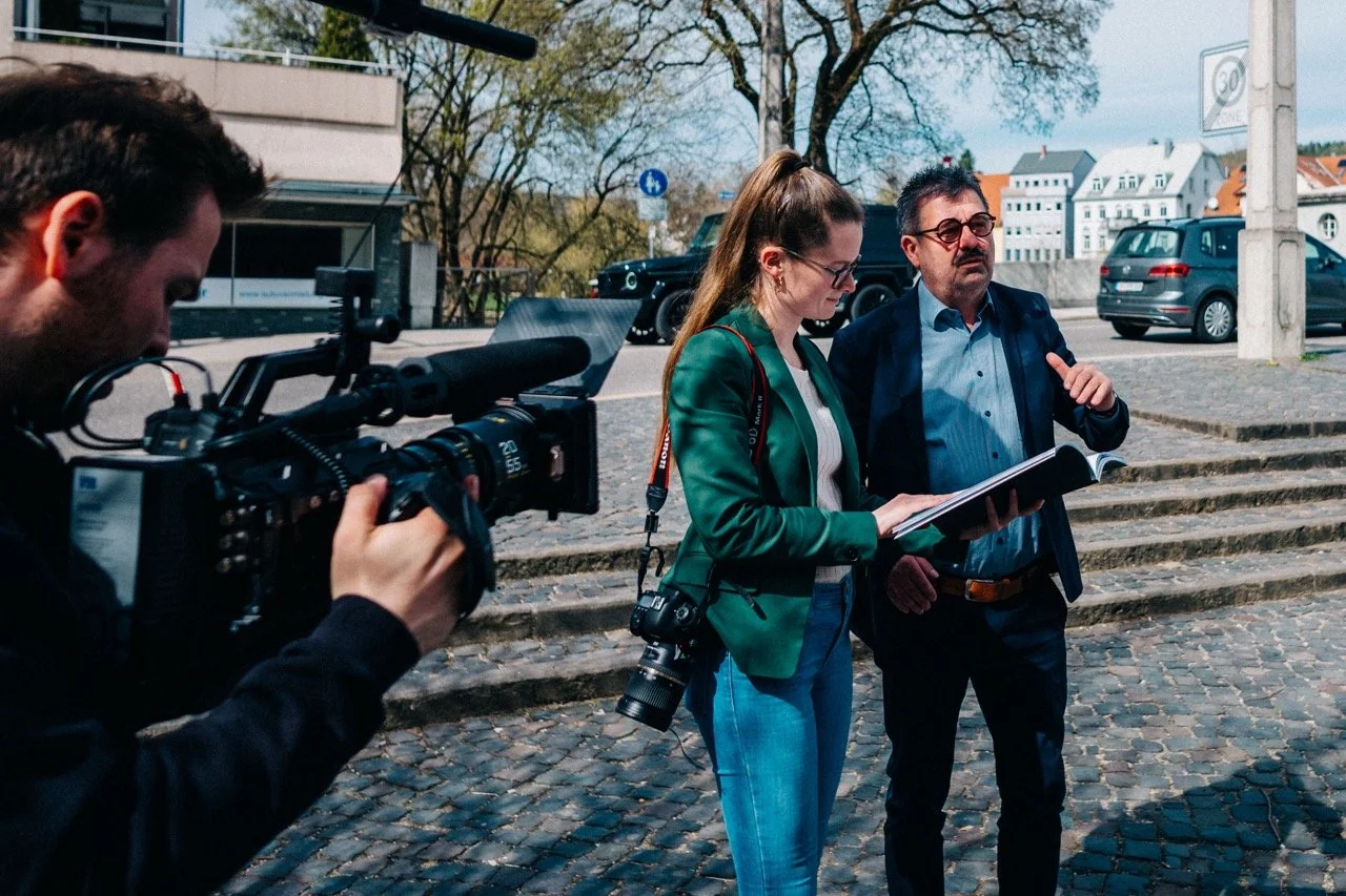 A woman and a man stand on a cobblestone street, reading documents, while a cameraman films them.