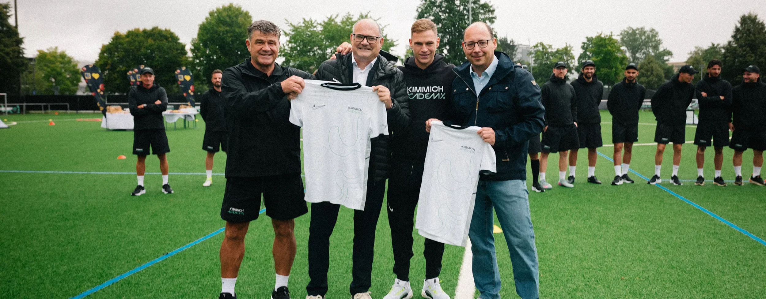 Four men standing on a football field holding signed white jerseys, with a line of players and trees in the background.