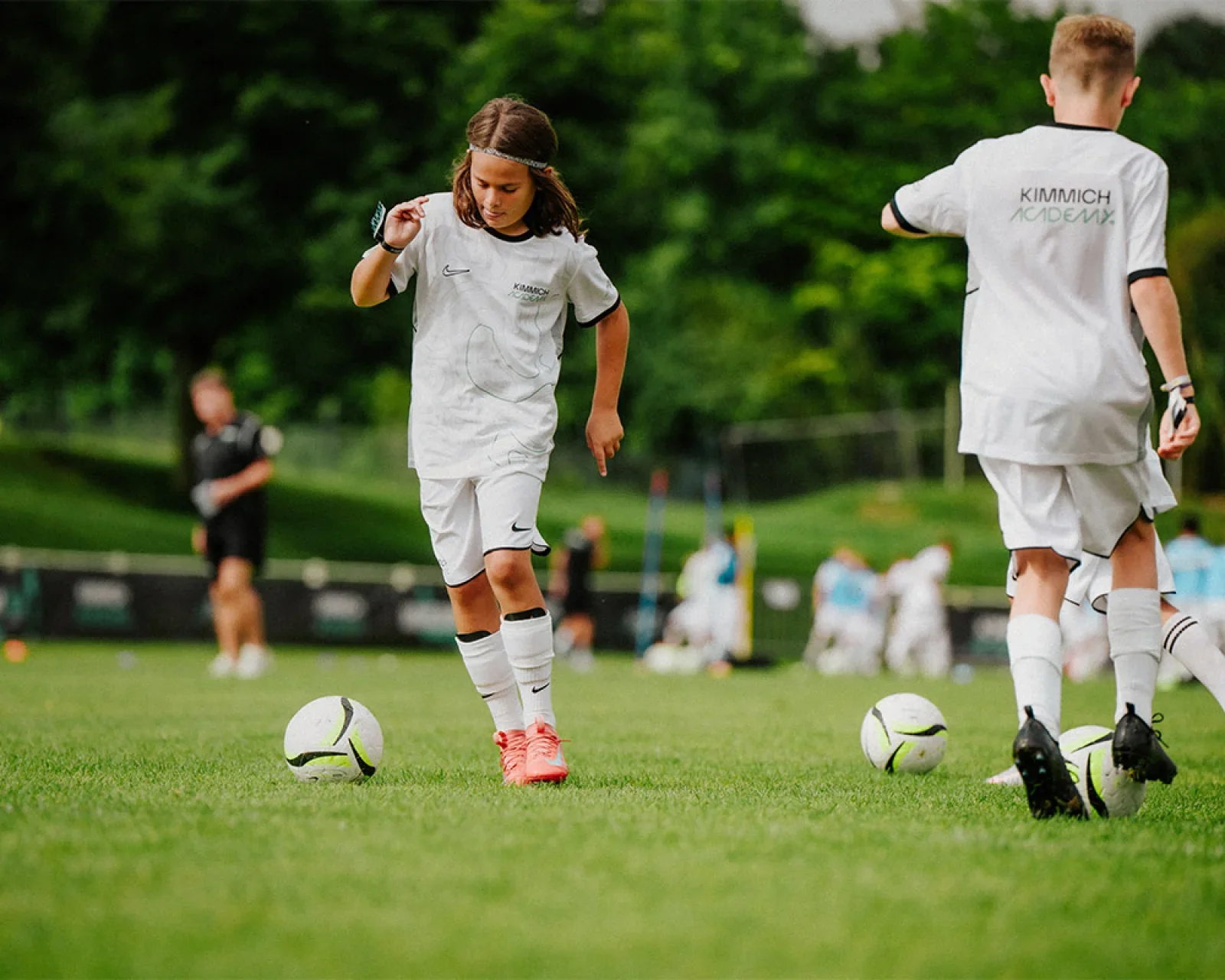 Children in white soccer uniforms practicing on a grassy field with soccer balls.