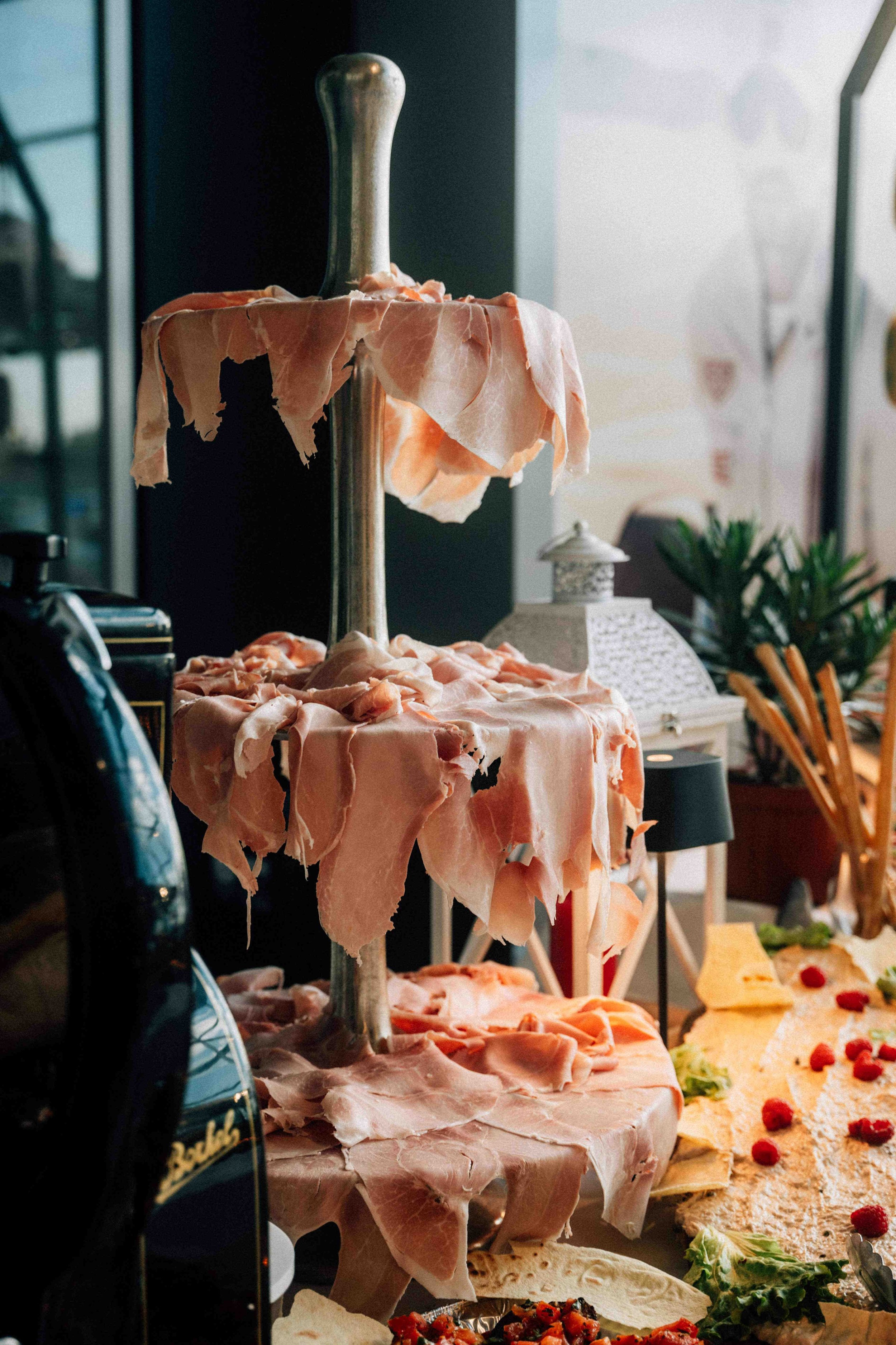 A food display with slices of ham or prosciutto draped over a tall metal stand, with other food items and decorations visible in the background.