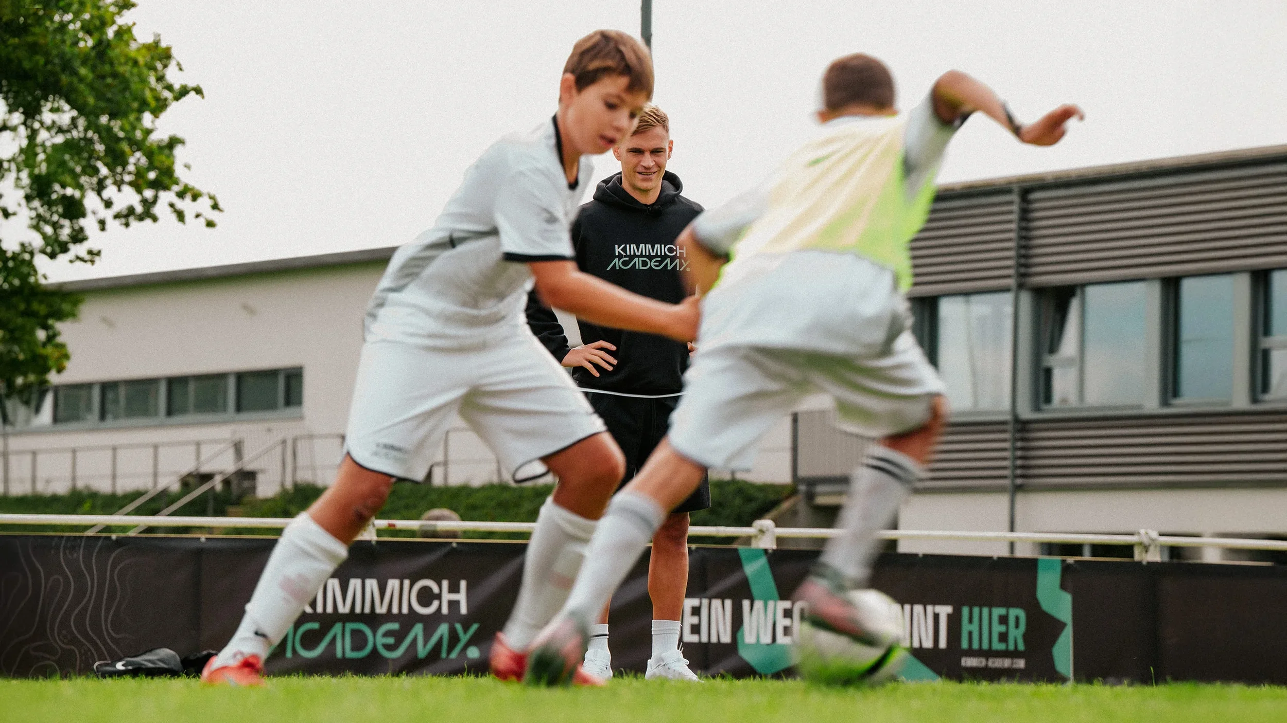 Two young soccer players in white uniforms compete for the ball while a coach watches in the background during a practice or game on a grassy field outside.