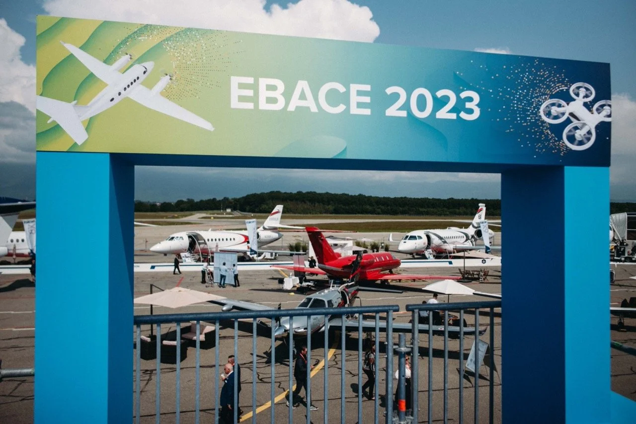 EBACE 2023 aviation exhibition with several private jets on display, a sign with a drone and airplane graphic, and a group of people walking near the aircraft.