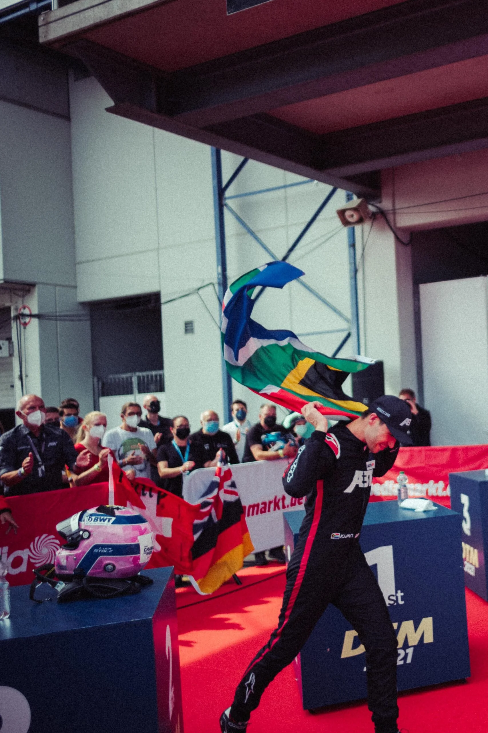 A Formula 1 driver holding South African flag at victory podium, with racing helmet on a table nearby, and spectators in the background wearing masks at a motorsport event.