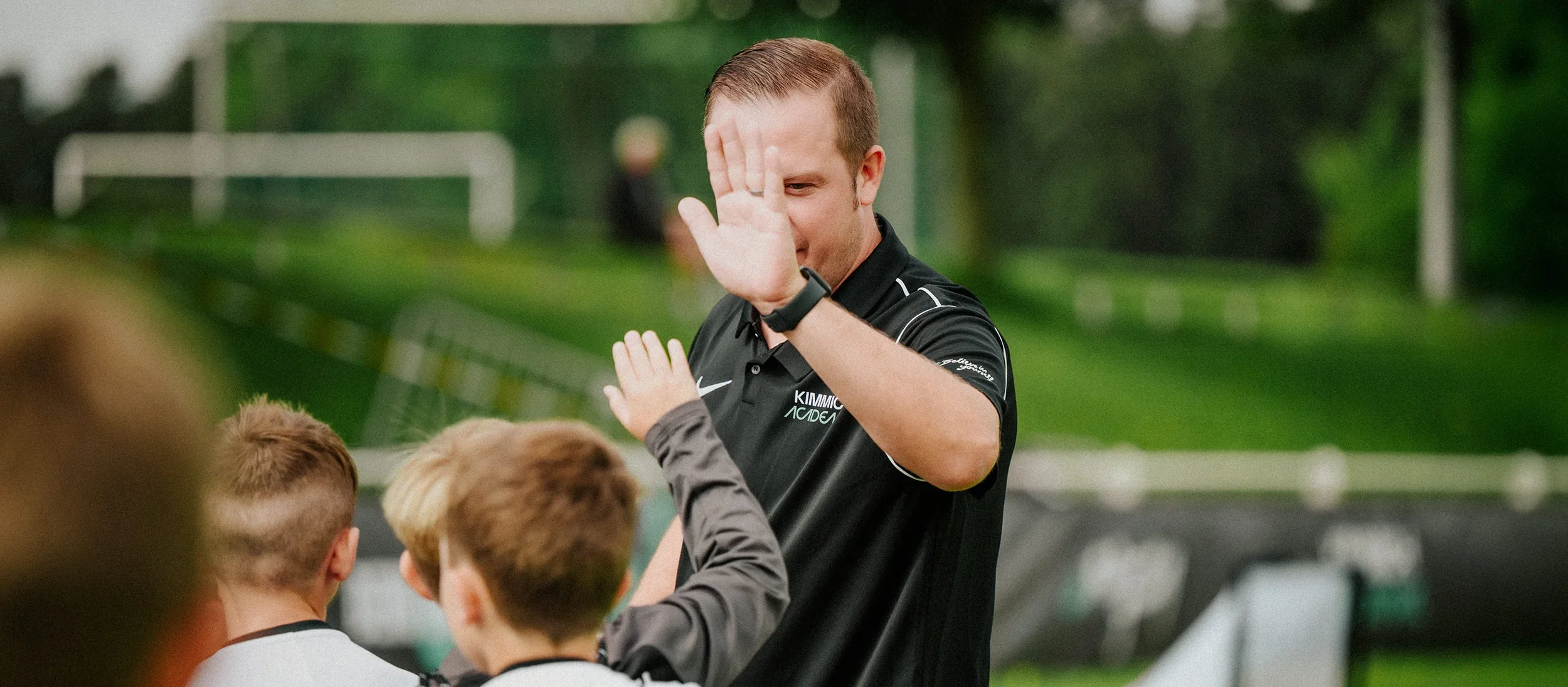 A man with short brown hair giving a high five to children at an outdoor sports field.