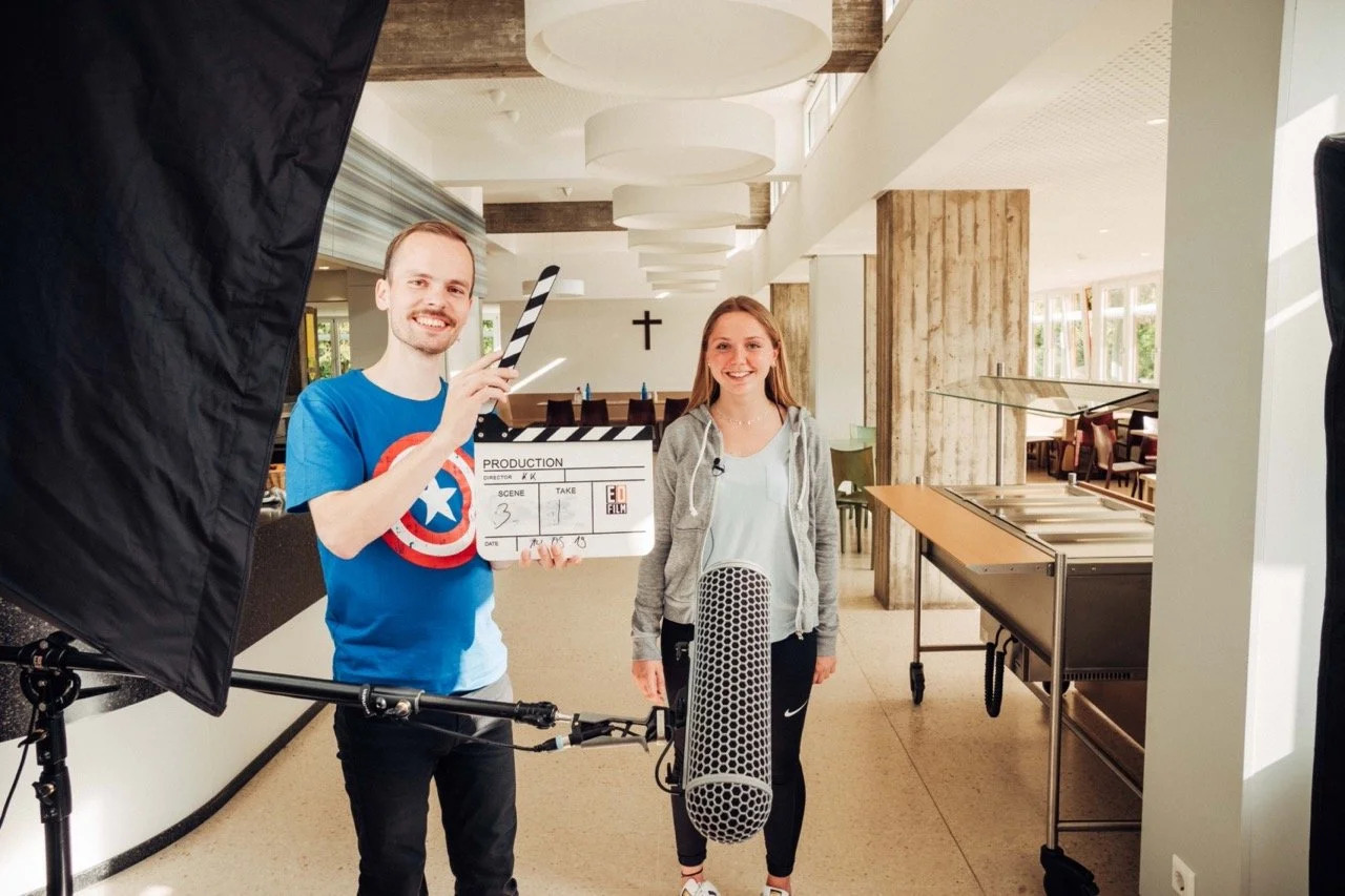 A man holding a clapperboard and a woman standing in a well-lit indoor space, possibly a dining area or lobby, with filming equipment visible in the frame.
