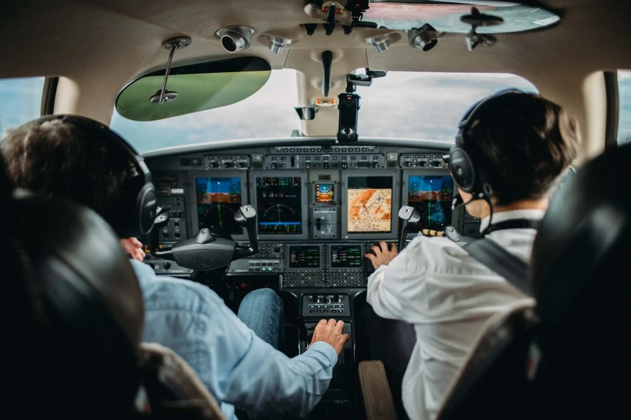 Inside the cockpit of an airplane with two pilots wearing headsets, looking at their instruments and screens during flight.