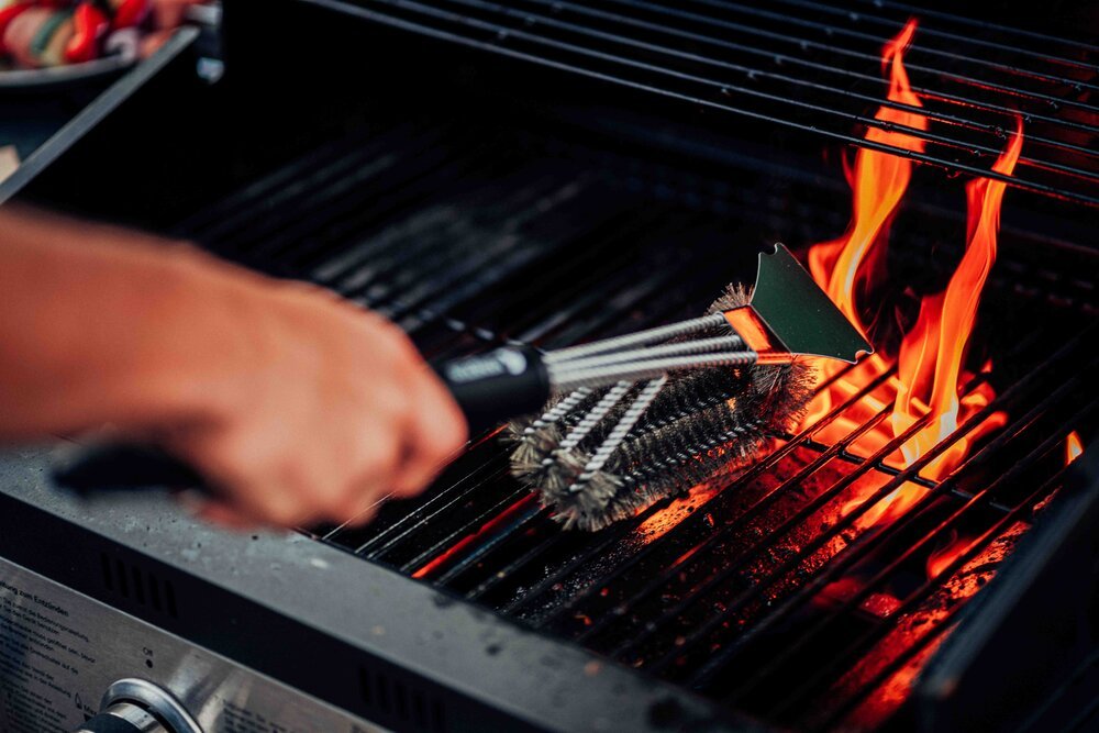 Person cleaning a grill with a wire brush while flames are visible on the barbecue.