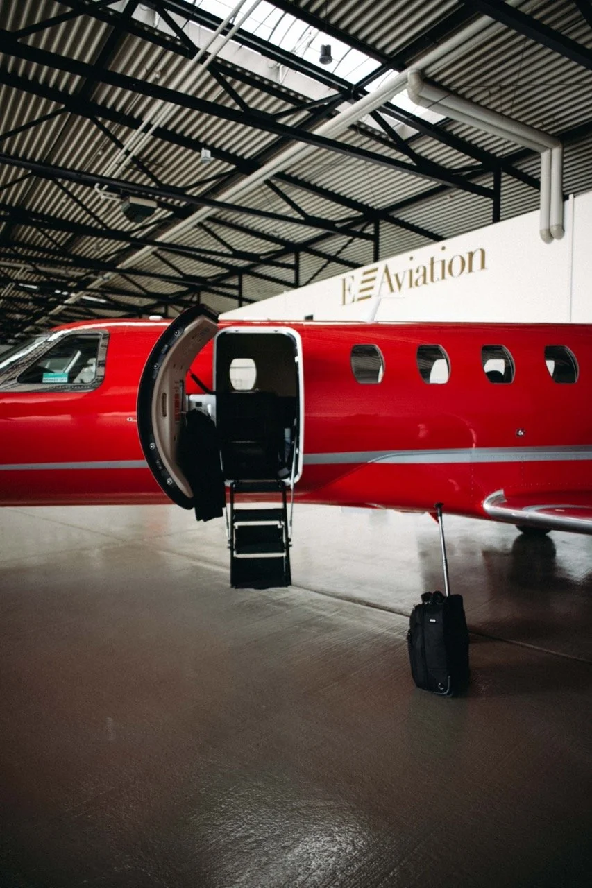 A red private jet with an open door and a small staircase leading inside, located inside an airport hangar labeled 'Aviation', with a black carry-on suitcase and a small black bag on the ground near the plane.