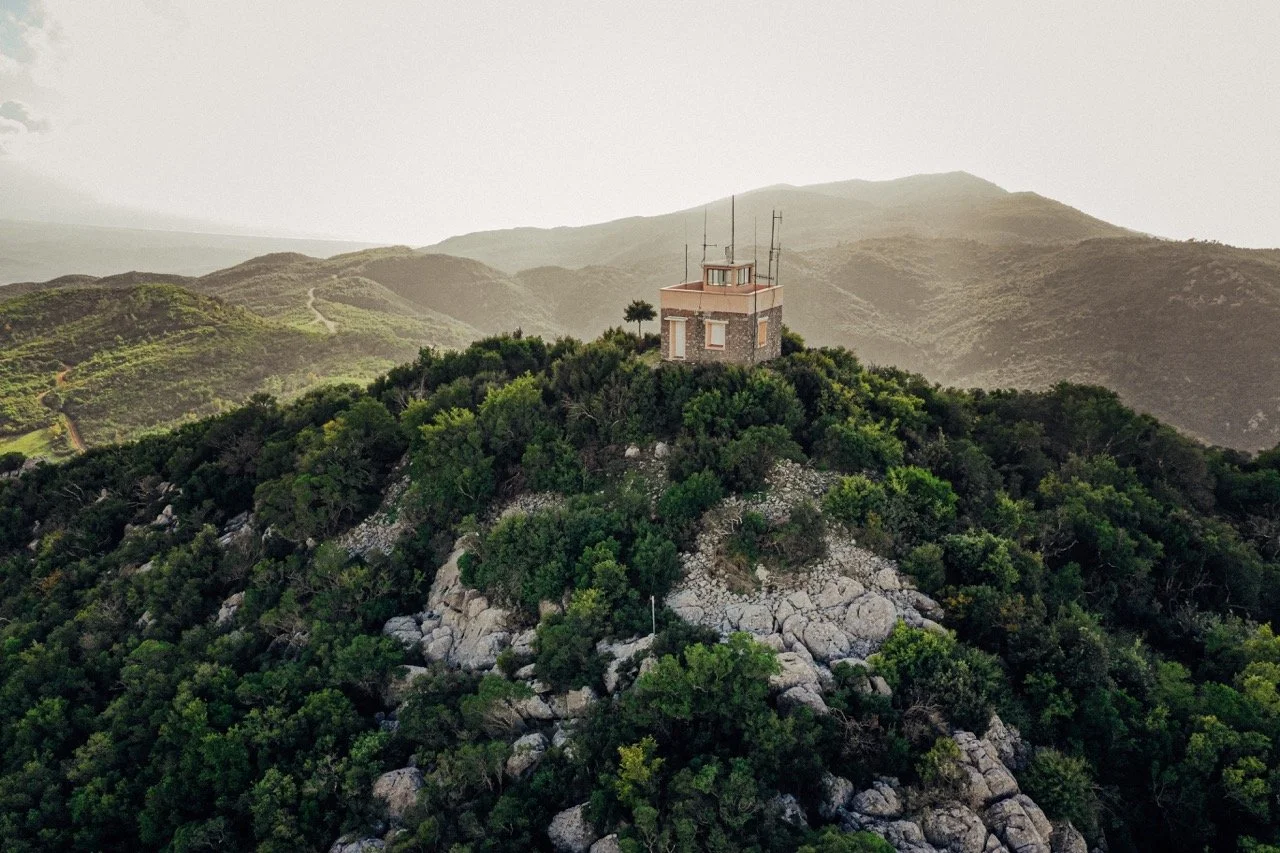 A small building with antennas on top, perched on a lush green hill with rocky terrain, surrounded by rolling hills and mountains in the background.