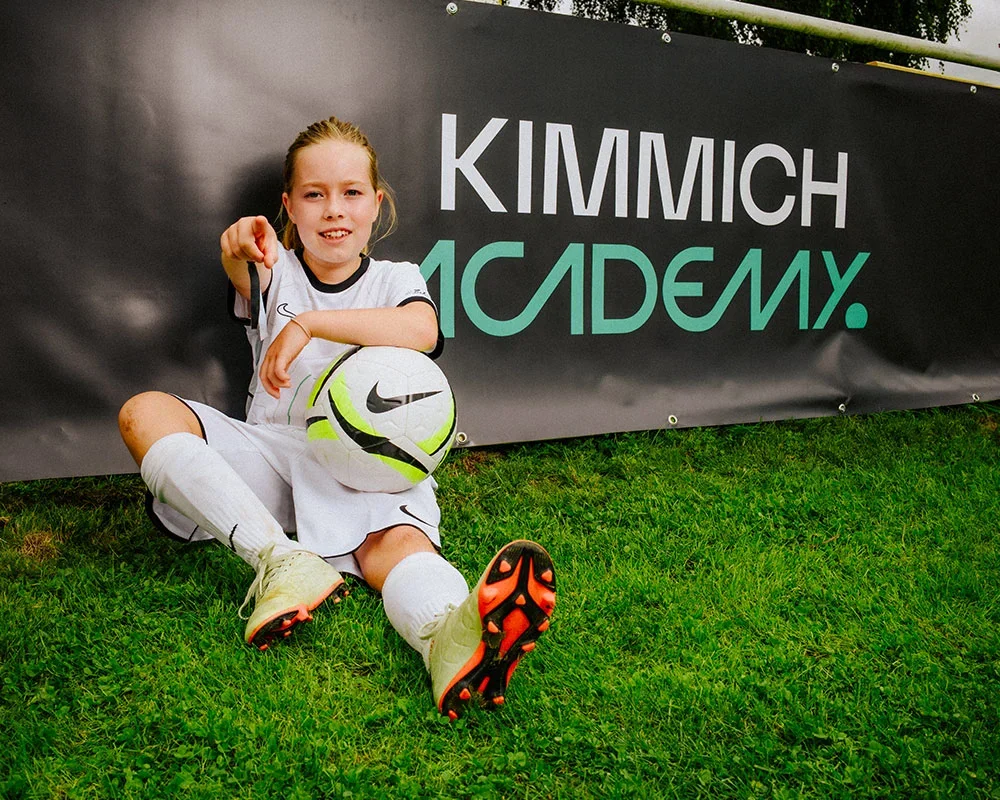 A young girl in a soccer uniform sitting on the grass with her soccer ball, smiling at the camera, near a Kimmich Academy banner.