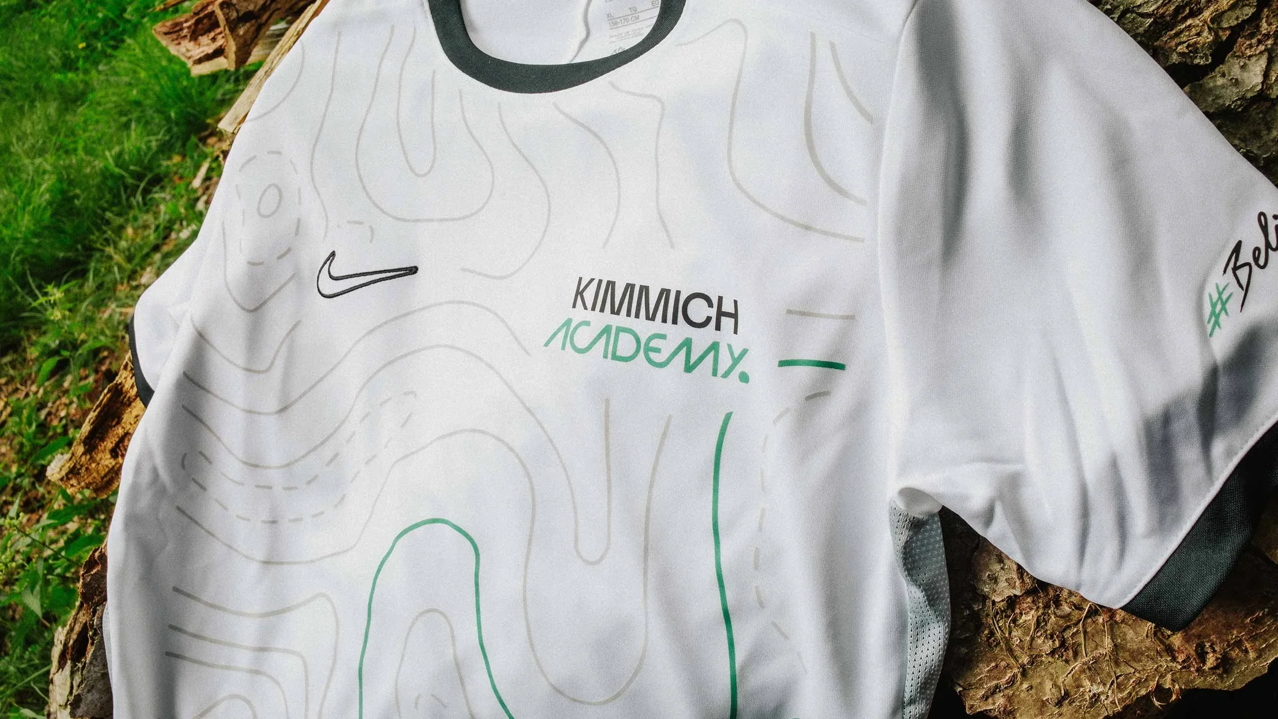 White sports jersey with black, green, and gray accents, lying on tree bark and grass, featuring the Nike arrow logo, and text including 'KIMMICH ACADEMY' and '# Be'.
