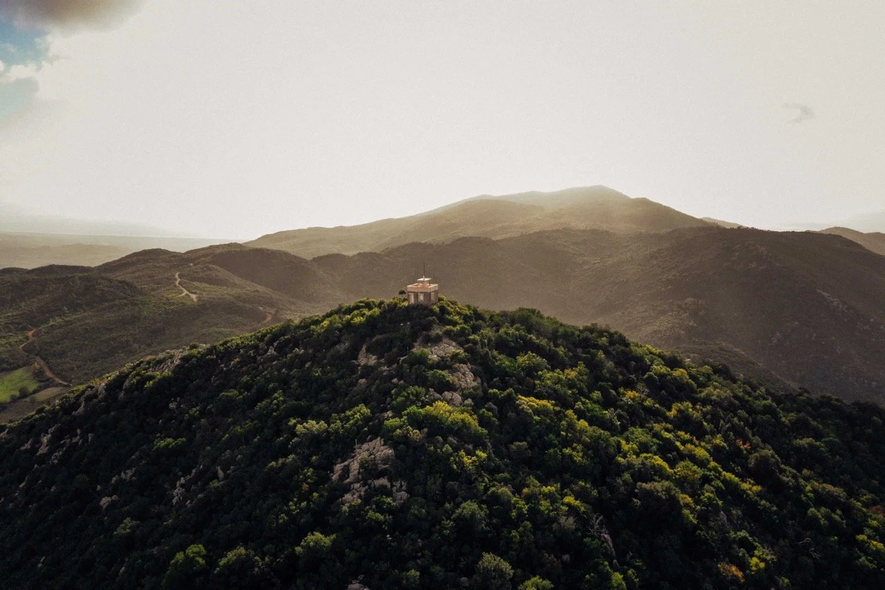A small building is situated on top of a lush green hill, with mountain ridges in the background under a cloudy sky.