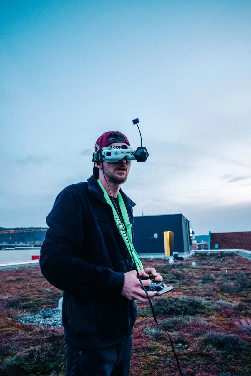 A man using a drone controller outdoors at dusk, wearing a head-mounted display and protective gear for drone flying.