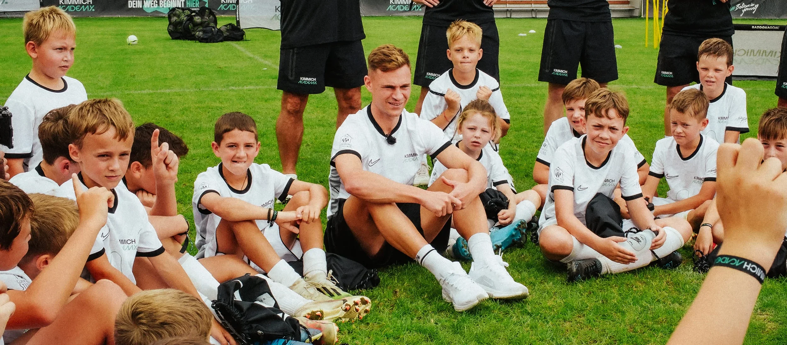 A group of young children and a coach sitting on the grass after a soccer practice or game, some are raising their fists and some are smiling, with soccer gear around them.