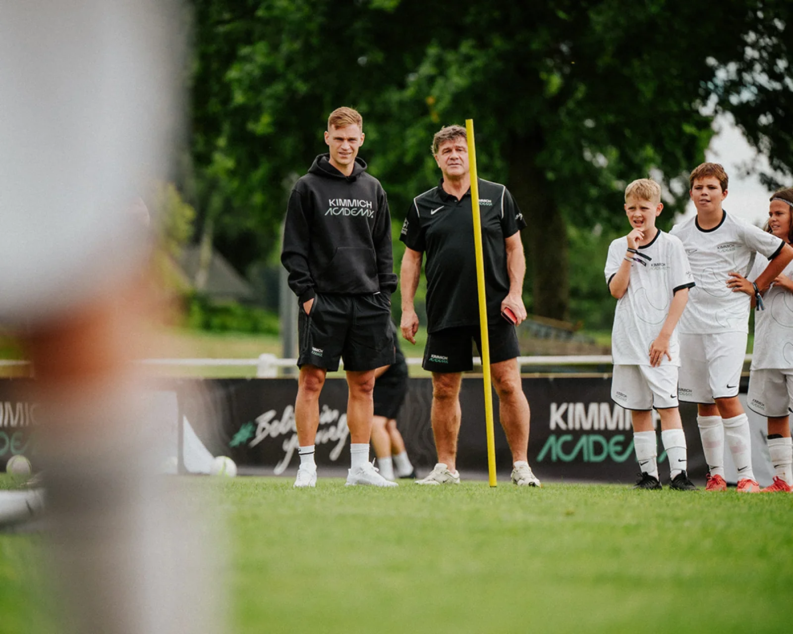 Soccer players and coach standing on a field during a practice or game, with spectators in the background.